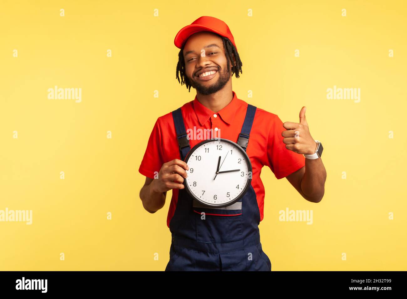 Joyeux messager ponctuel dans l'horloge uniforme et montrant les pouces vers le haut, comme geste, satisfait avec un excellent service de livraison à l'heure.Studio d'intérieur isolé sur fond jaune. Banque D'Images