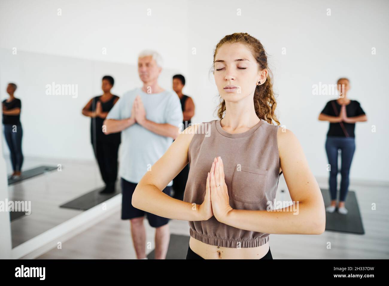 Une jeune femme avec les yeux fermés a joint ses mains méditant en cours de yoga avec d'autres personnes en arrière-plan Banque D'Images