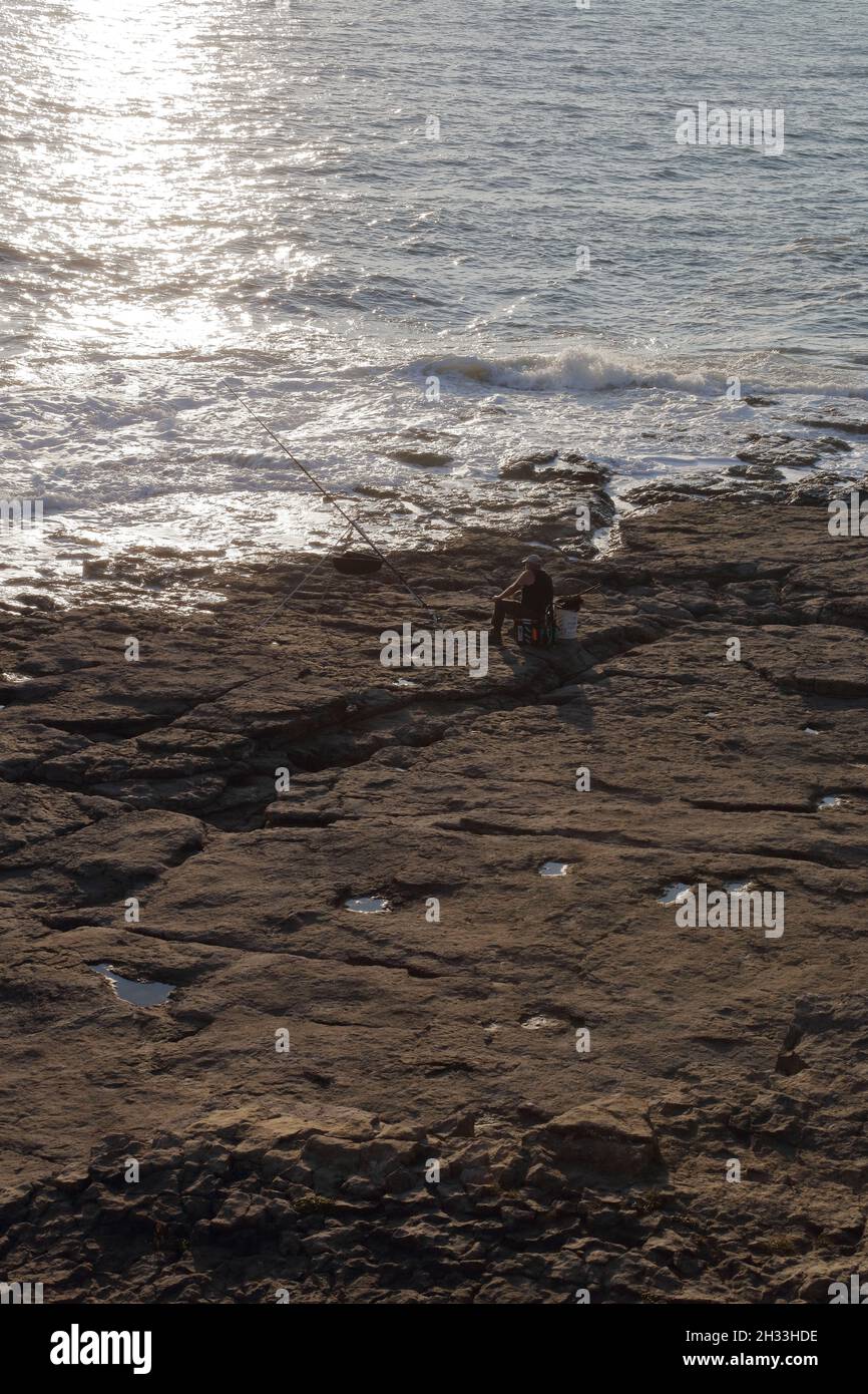 Une journée de pêche, s'assit patiemment en attendant ce remorqueur toujours insaisissable sur la ligne juste au-dessus des eaux qui s'écrasent sur les rochers. Banque D'Images