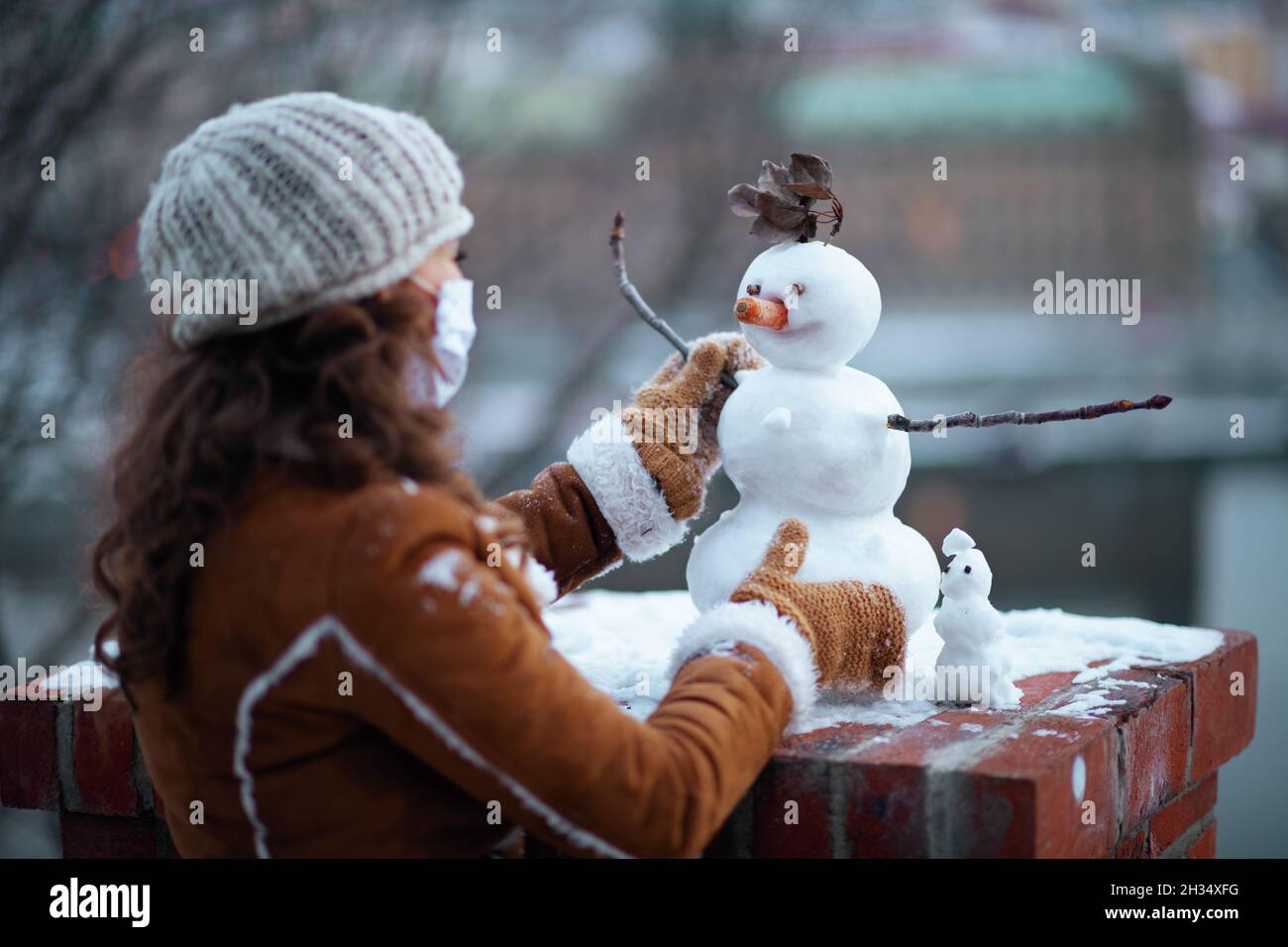 Vue de derrière une femelle avec des mitaines et un masque médical dans un bonnet tricoté et un manteau de peau de mouton faisant un bonhomme de neige dehors dans le parc de la ville en hiver. Banque D'Images