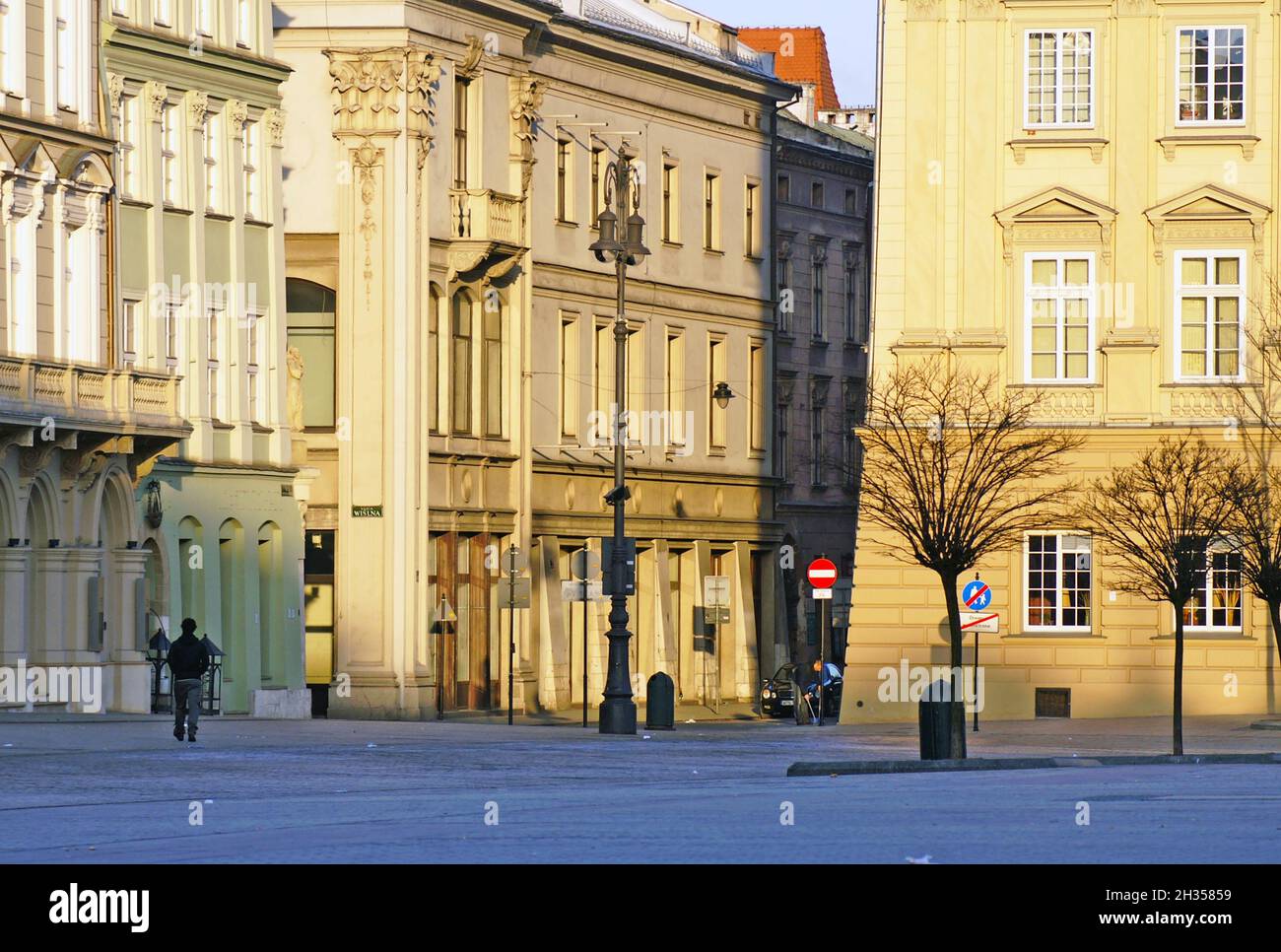 Le lever du soleil illumine l'architecture d'Europe de l'est, qui fait partie de la plus grande place médiévale d'Europe à Cracovie, en Pologne.Le froid matin d'hiver peut être vu dans la personne de marche groupée et les arbres sans feuilles signifiant la saison d'hiver. Banque D'Images