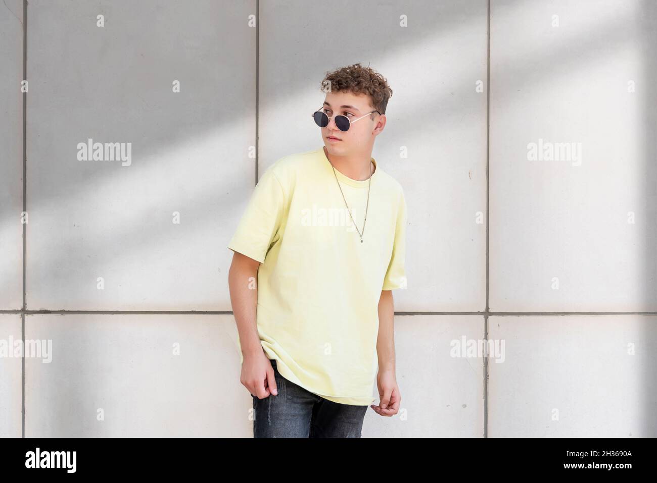 Jeune homme en t-shirt jaune posant avec ses lunettes de soleil sur un fond en béton gris à l'extérieur Banque D'Images