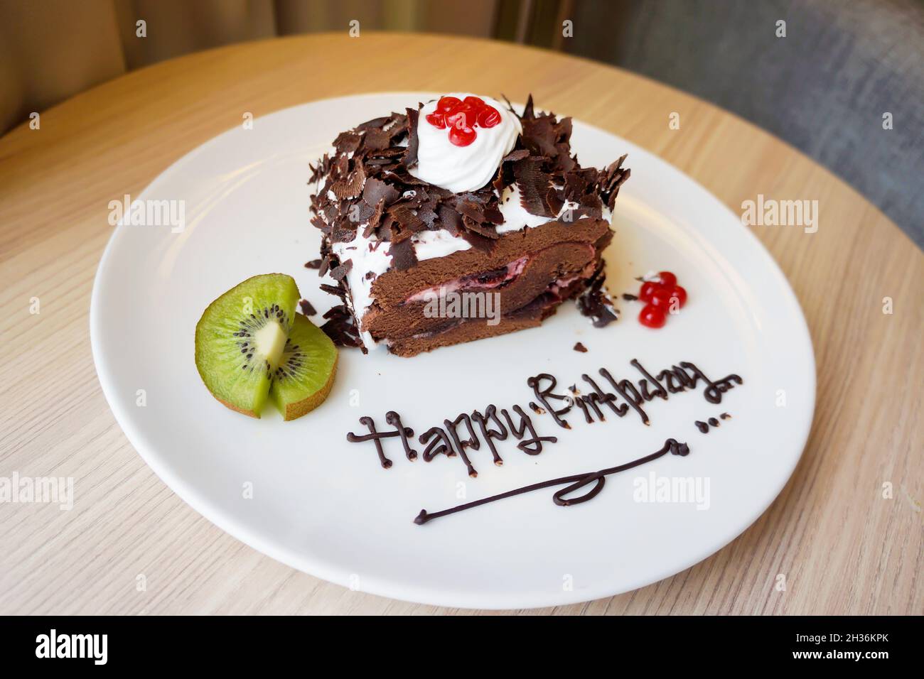 Vue rapprochée du gâteau d'anniversaire au chocolat avec crème fouettée, kiwi en tranches et cerise sur une assiette blanche à la table.Arrière-plan bokeh.Personne. Banque D'Images