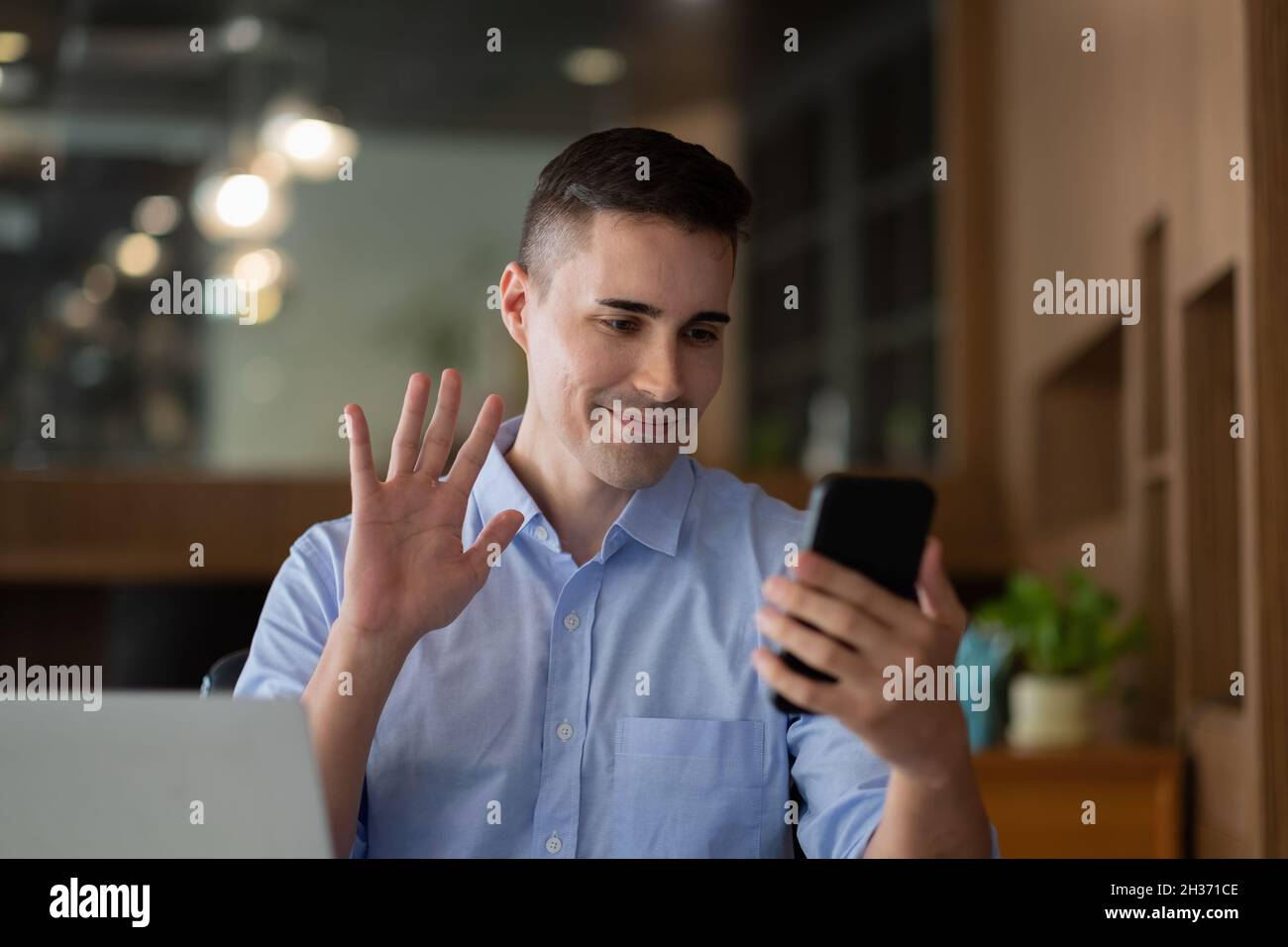 Homme d'affaires joyeux et joyeux utilisant l'application pour smartphone pour la communication virtuelle, tenant le téléphone à la main et en agitant à la webcam, tenant des appels vidéo assis à Banque D'Images