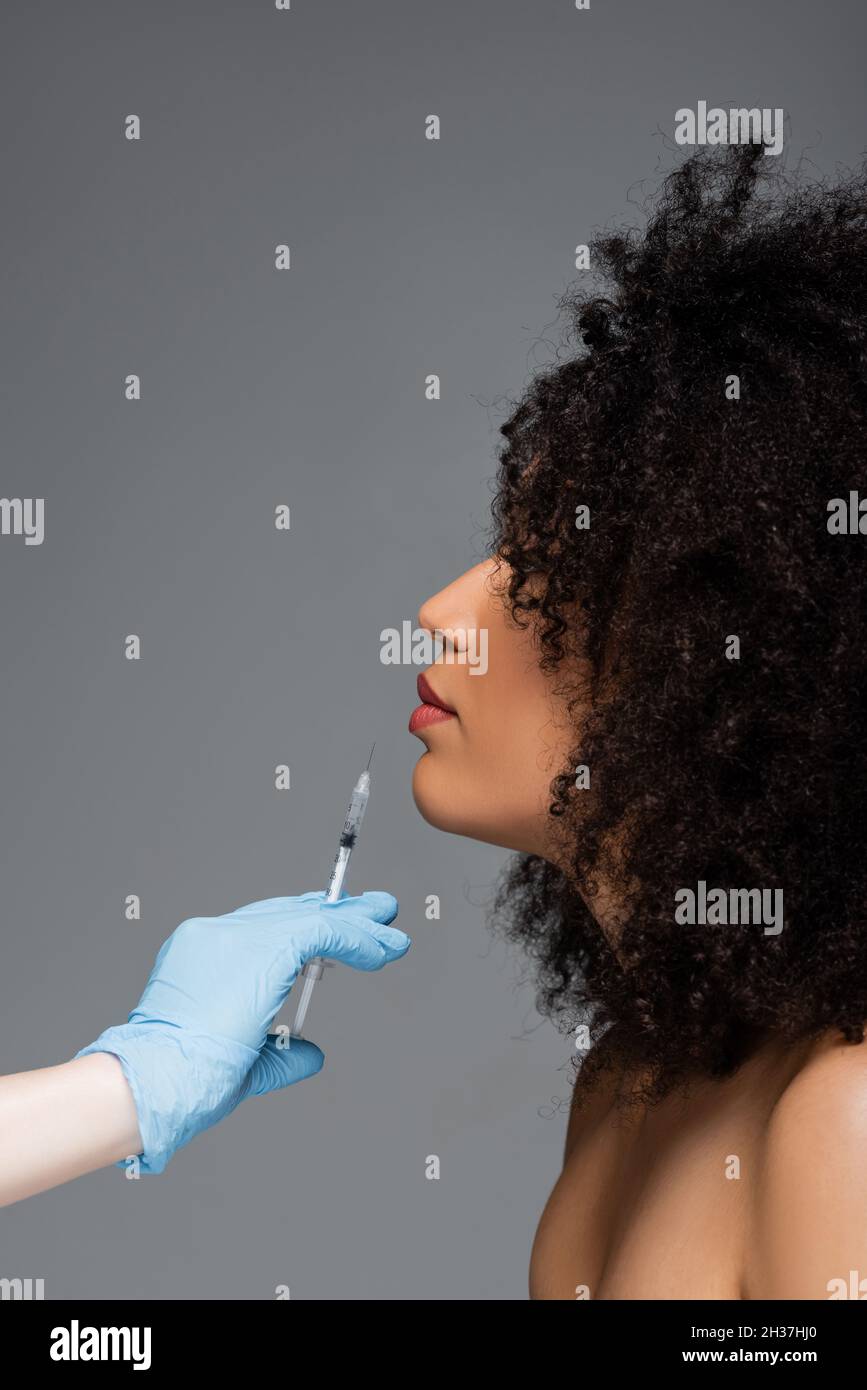 vue latérale de la femme afro-américaine près de cosmetologiste avec injection de beauté isolée sur gris Banque D'Images