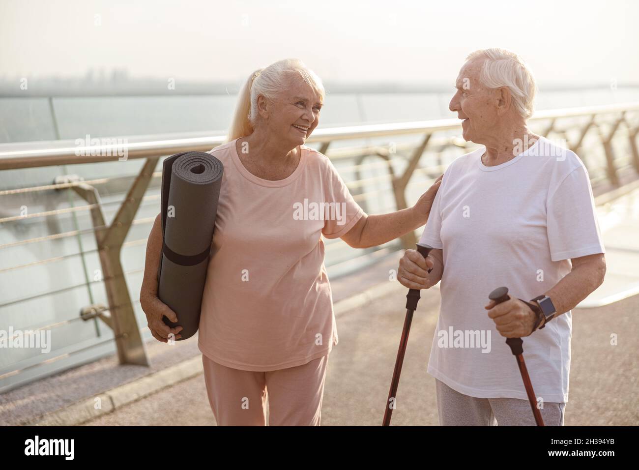 Une femme aîée souriante avec tapis salue un homme avec des bâtons pour la marche nordique sur la passerelle Banque D'Images