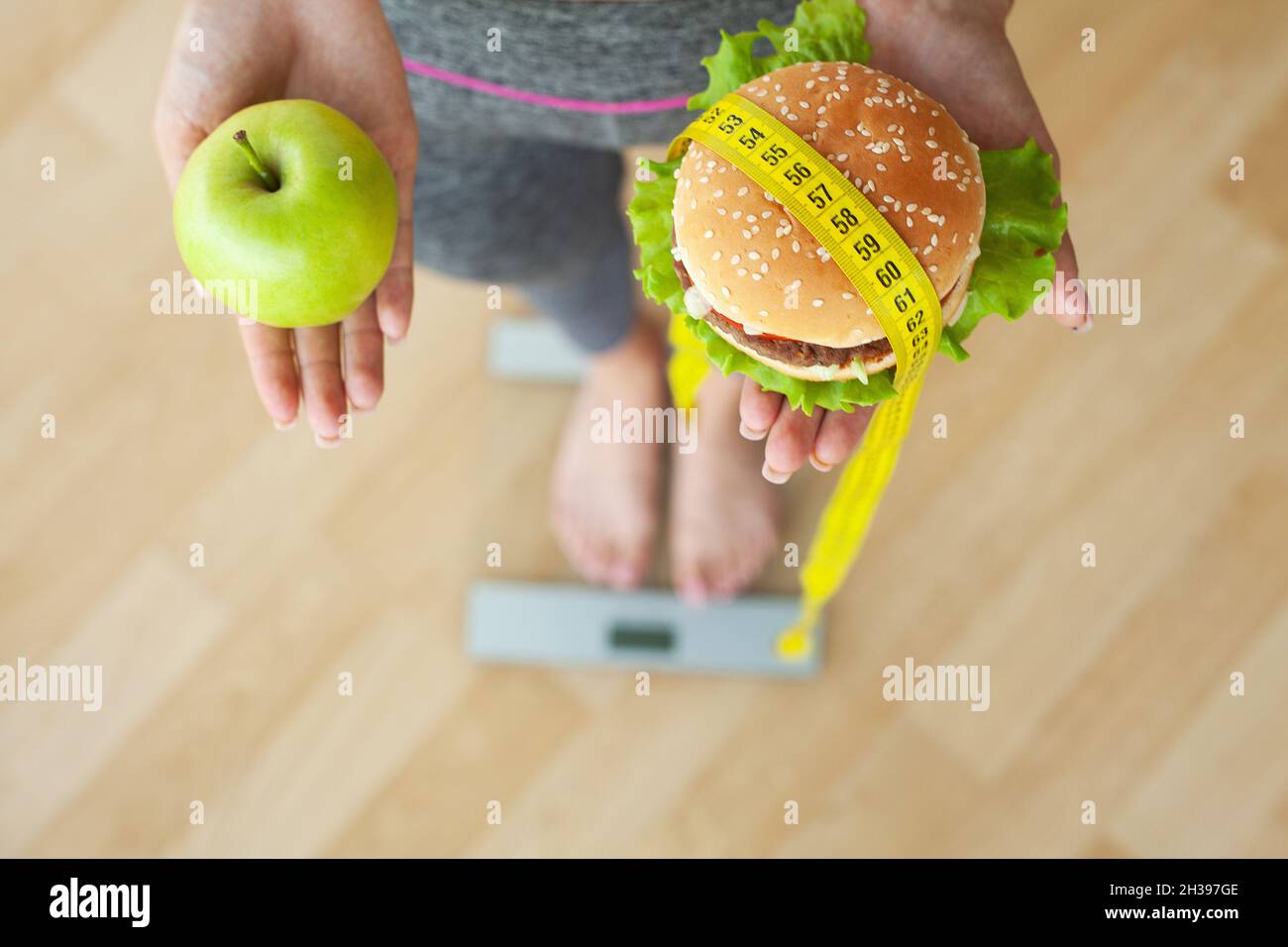 Jeune fille attrayante fait le choix entre la nourriture saine et nocive tenant la pomme et un hamburger dans ses mains. Banque D'Images