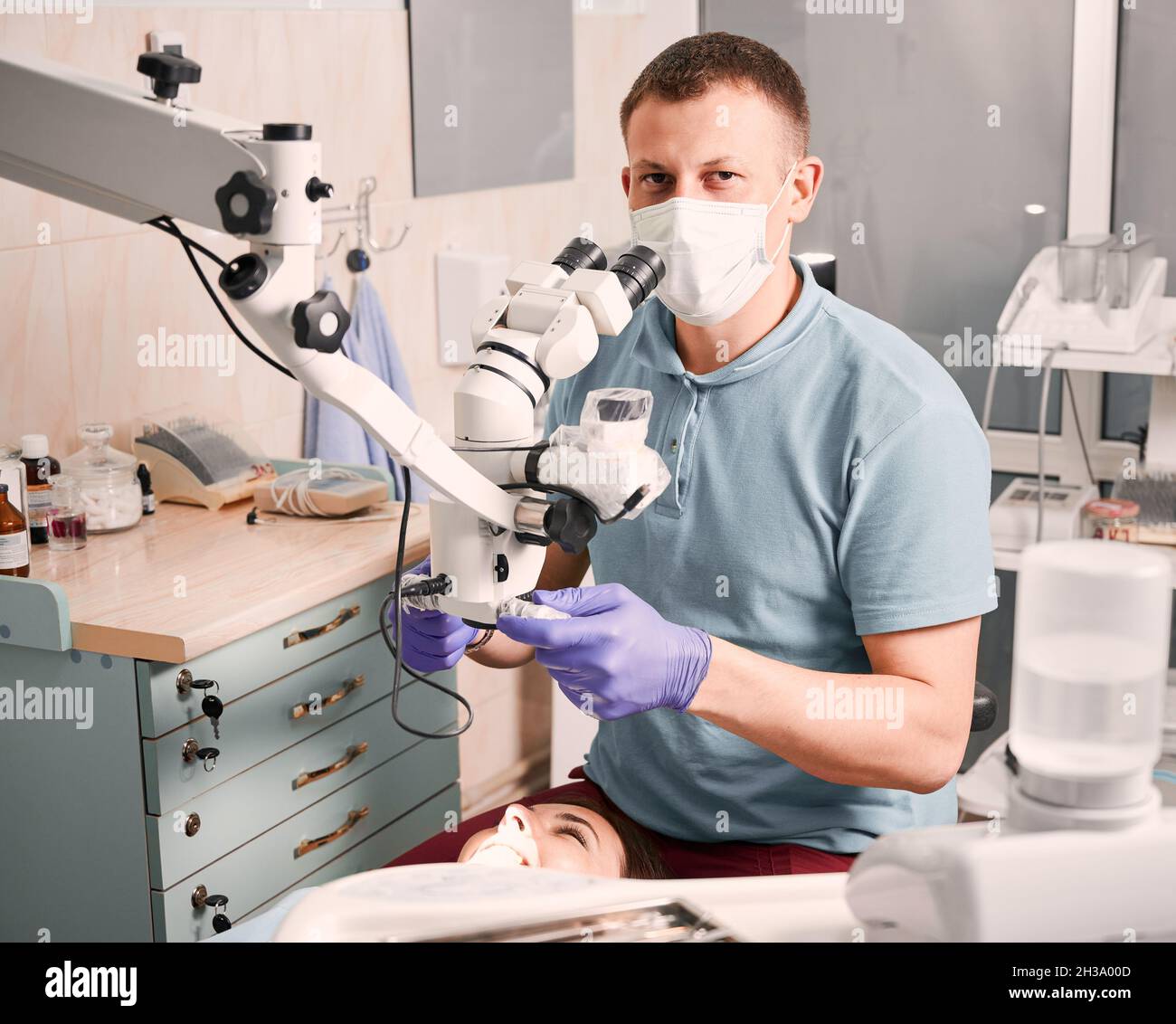 Stomatologiste mâle dans des gants stériles utilisant un microscope de diagnostic dentaire alors que la patiente est allongée dans une chaise dentaire.Jeune homme dentiste dans un masque médical regardant la caméra tout en étant assis à côté de la jeune femme Banque D'Images