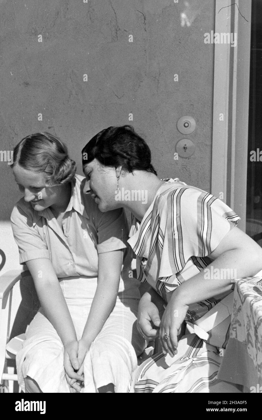 Zwei Frauen, vor einem Haus in der sonne sitzend, schauen zu ihrer rechten Seite und lächeln; Deutschland 1930er Jahre.Deux femmes, assises devant une maison au soleil, regardant de leur côté droit et souriant; Allemagne des années 1930. Banque D'Images