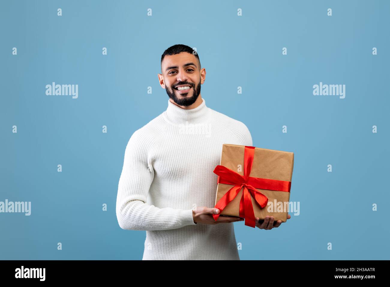 Le meilleur cadeau pour vous.Joyeux jeune arabe tenant une boîte cadeau enveloppée, souriant à l'appareil photo, debout sur fond bleu Banque D'Images