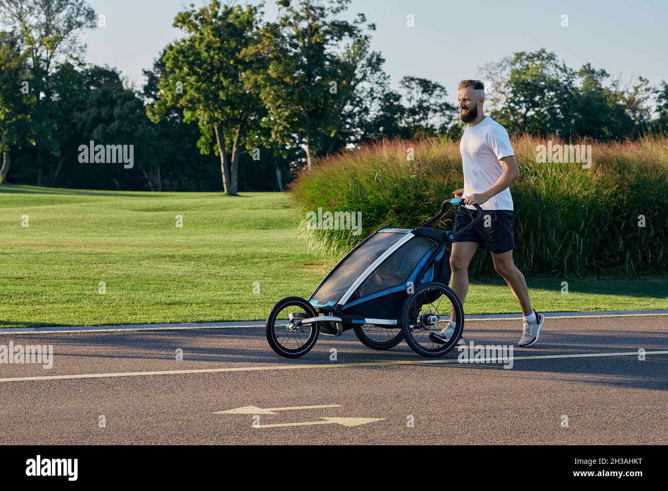homme sportif dans la course de vêtements de sport avec poussette de bébé au matin ensoleillé dans le parc de la ville pendant le temps de la paternité. Vue latérale Banque D'Images