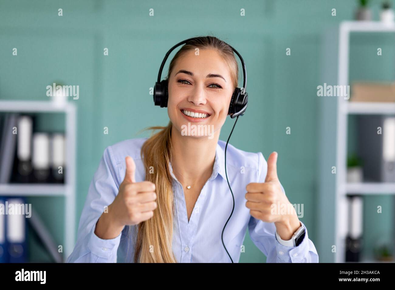 Enseignement à distance, travail.Bonne dame dans l'oreillette montrant le pouce vers le haut geste, regardant et souriant à l'appareil photo dans le bureau Banque D'Images