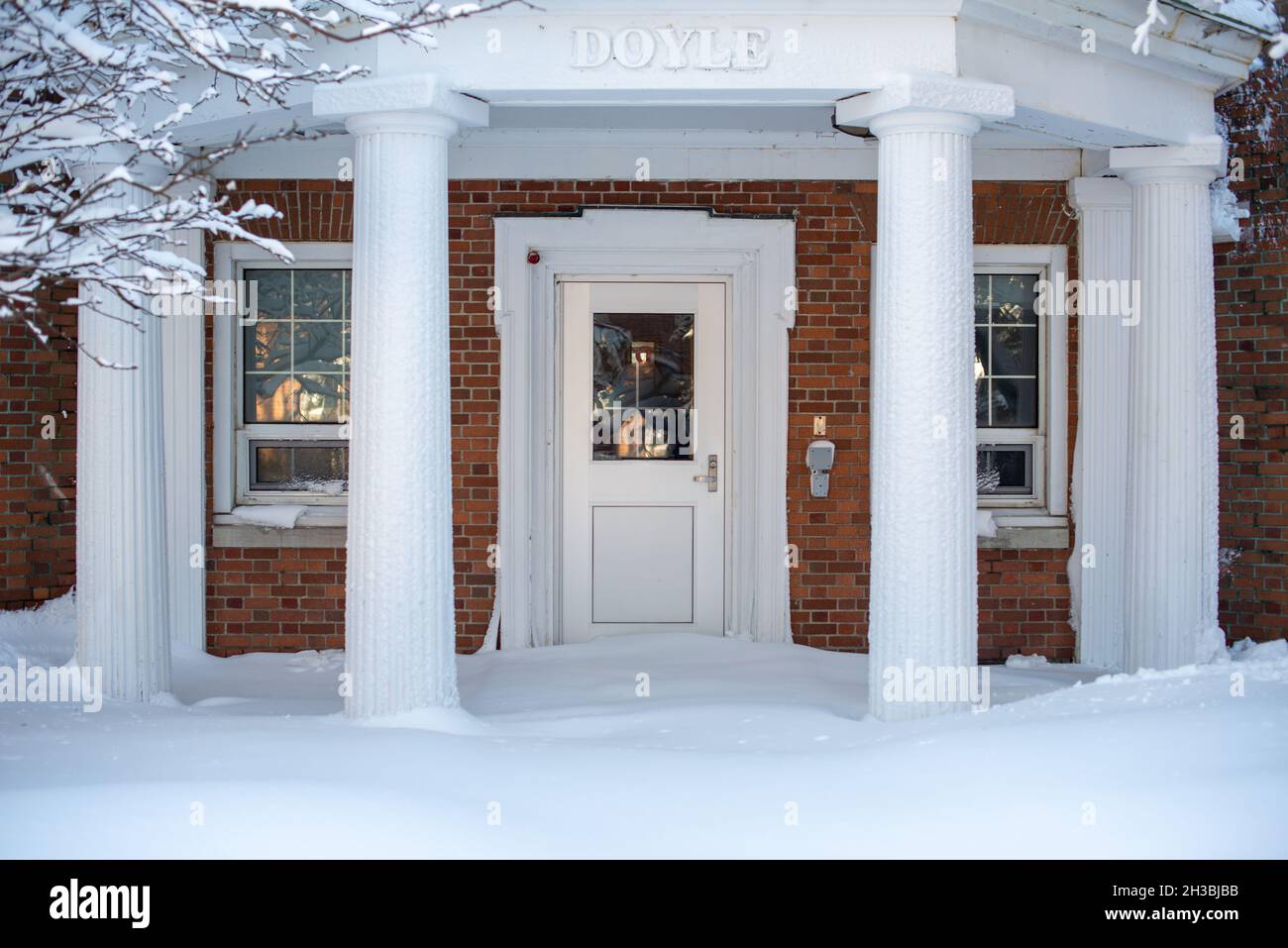 L'entrée extérieure d'un bâtiment en briques brunes avec une porte blanche, des colonnes, des arbres, des fenêtres et des snowdrifts bloque l'entrée ou la façade. Banque D'Images