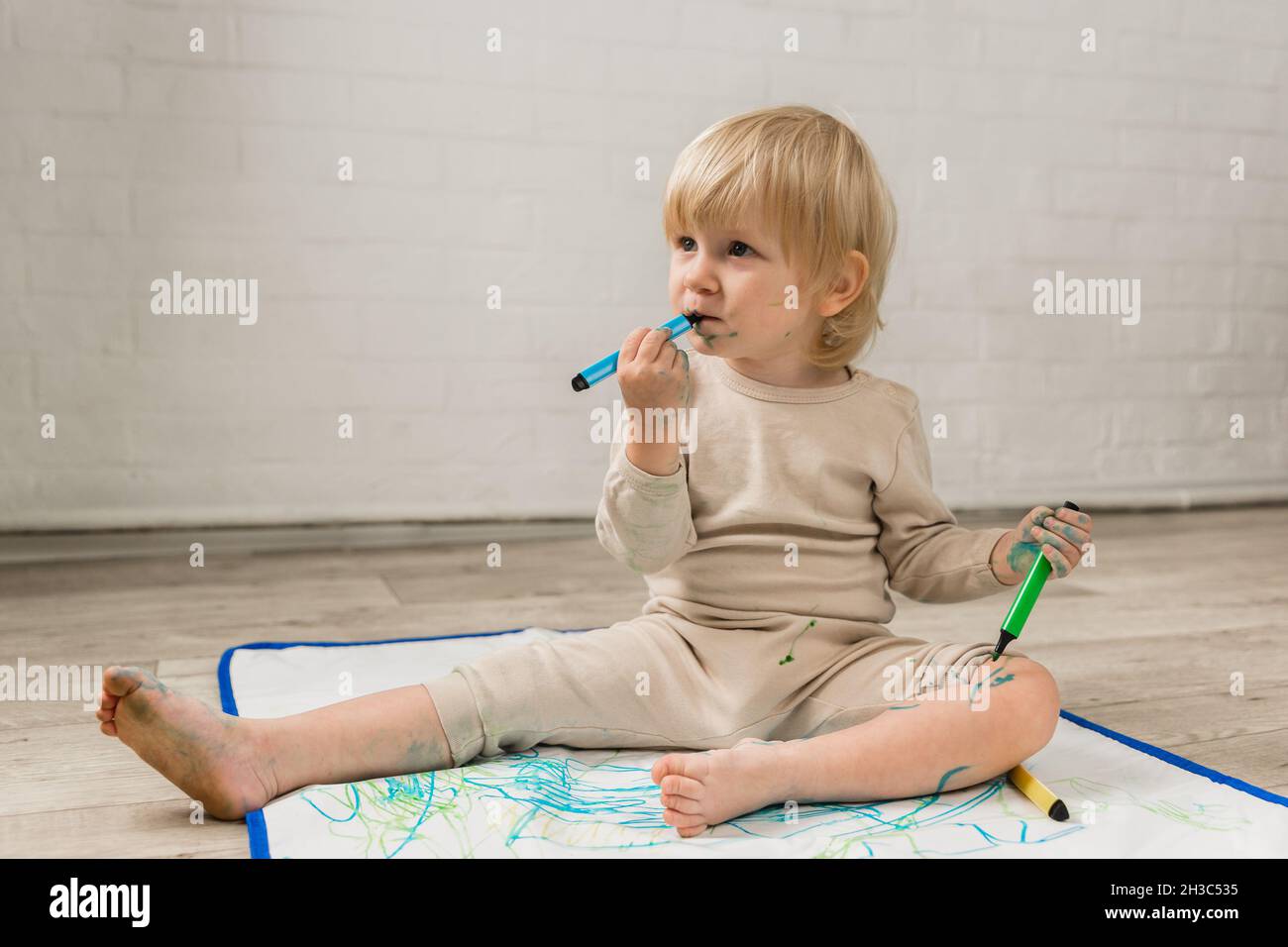 Une petite fille gaie en pyjama est assise sur le sol et dessin avec des marqueurs d'eau sur un chiffon spécial d'huile.Le visage et les vêtements sont tachés Banque D'Images