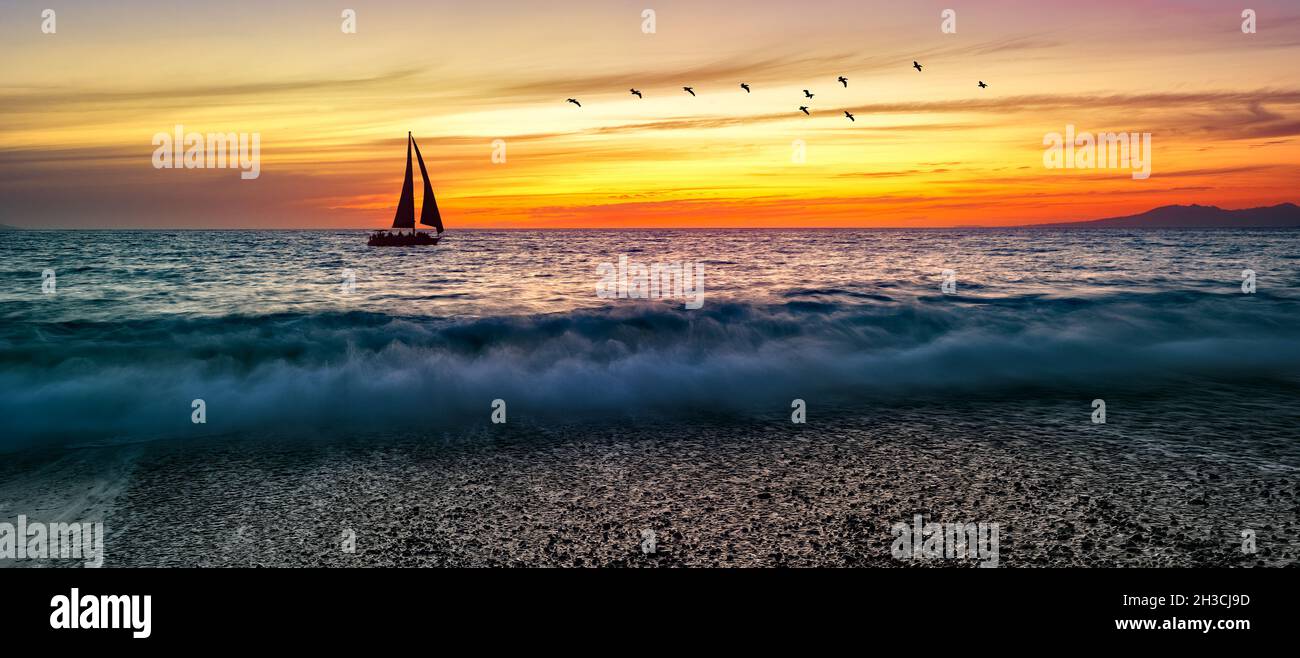 Un Voilier navigue le long de l'océan contre Un ciel coloré de coucher de soleil avec des oiseaux volant au format d'image Panorama Banque D'Images