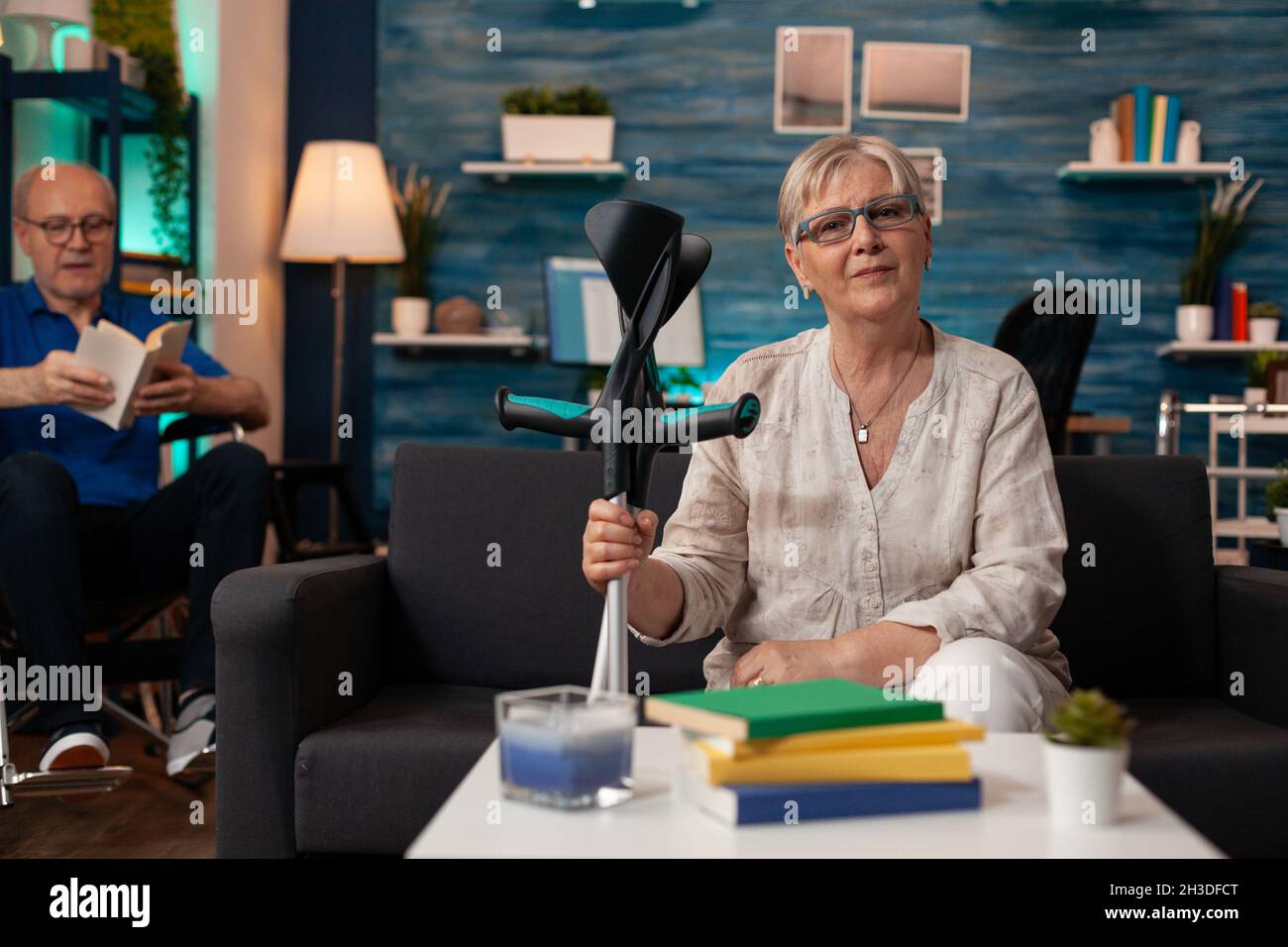 Vieille femme tenant des béquilles et regardant la caméra dans l'espace intérieur.Adulte âgé avec un handicap assis sur un canapé tandis que l'homme à la retraite lit en fauteuil roulant sur fond de salon Banque D'Images