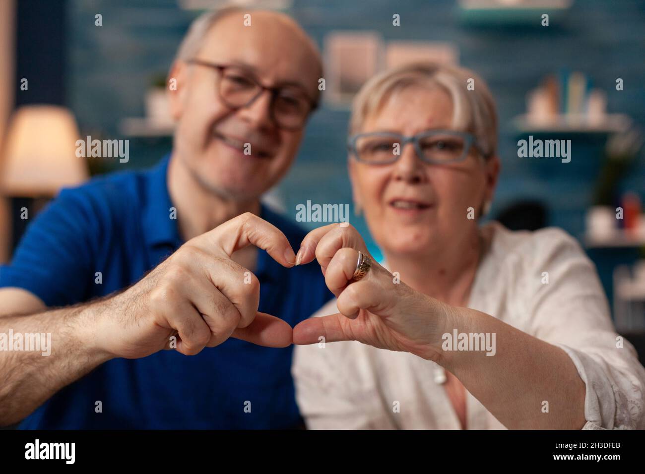 Couple aîné créant un signe de forme de coeur avec les mains à la maison.Des personnes âgées mariées dans l'amour faisant symbole romantique tout en regardant l'appareil photo dans le salon.Mari et femme montrant le chiffre d'affection Banque D'Images