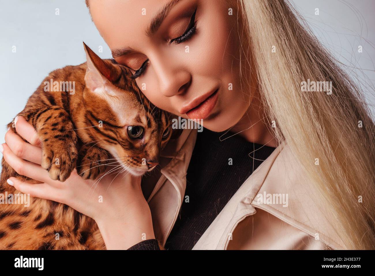Portrait d'une jolie jeune femme blonde avec un chat bengale.Belle fille, regarde le chat, coiffure élégante, maquillage impudent, blanc Banque D'Images