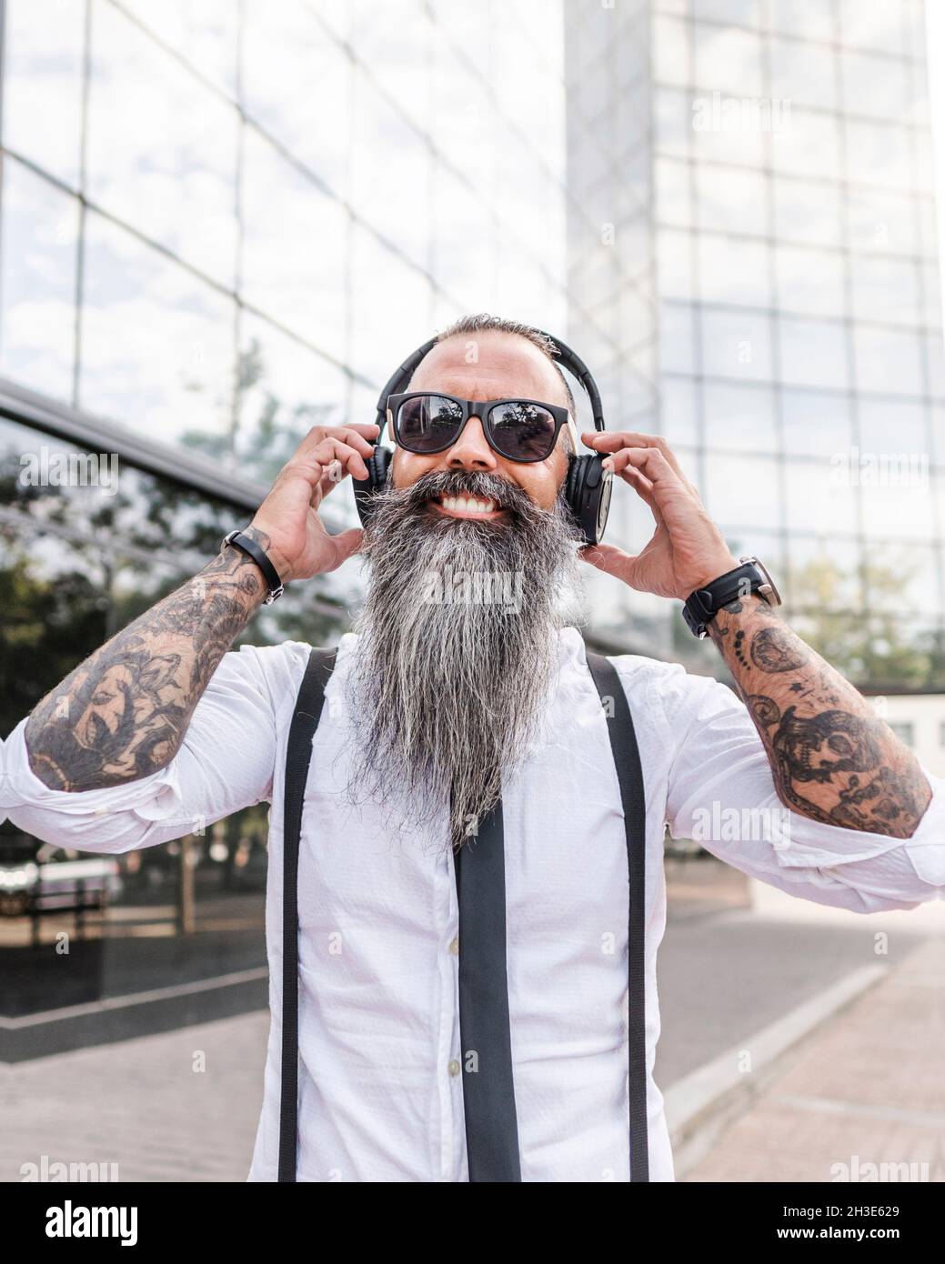 Happy Bearded hipster mâle en tenue habillée et lunettes de soleil écouter  de la musique avec des écouteurs sans fil tout en se tenant près des  bâtiments modernes Photo Stock - Alamy