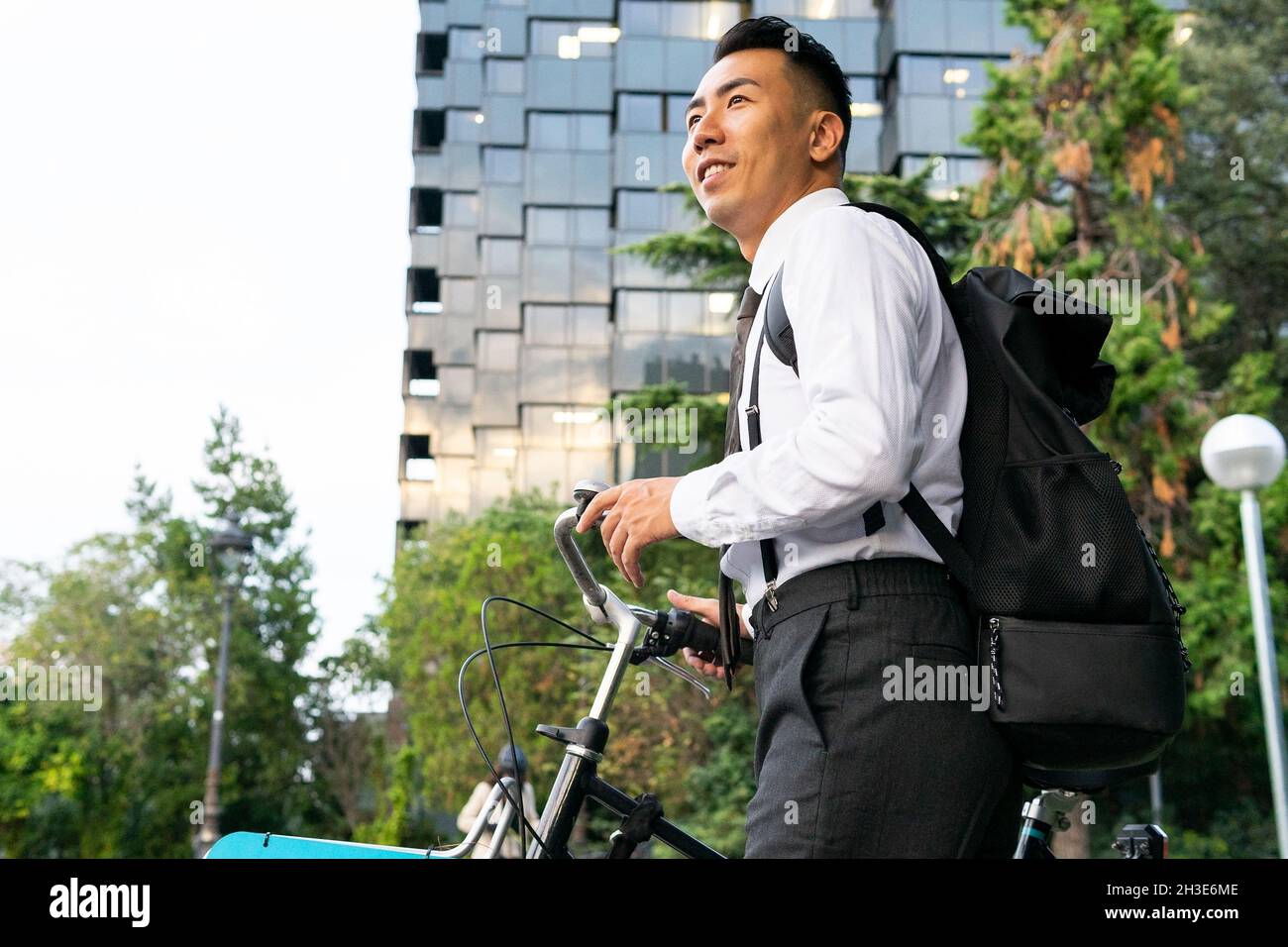 Vue latérale d'un jeune homme de bureau de rêve avec sac à dos et vélo, à l'écart des bâtiments urbains et des arbres Banque D'Images