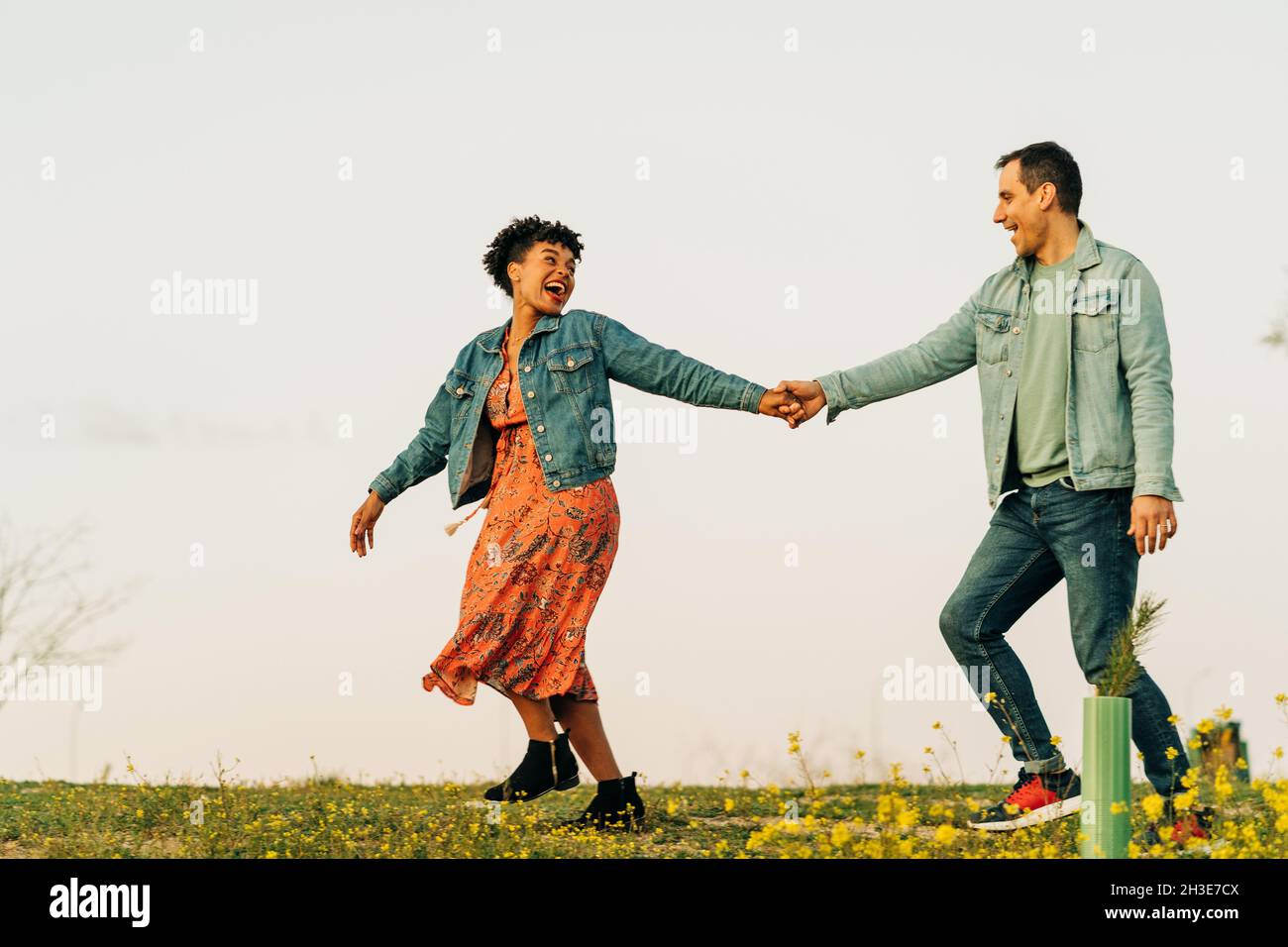 Pleine longueur de jeune positif multiethnique couple dans des vêtements décontractés tenant les mains et souriant tout en marchant sur la prairie pendant la date romantique en co Banque D'Images