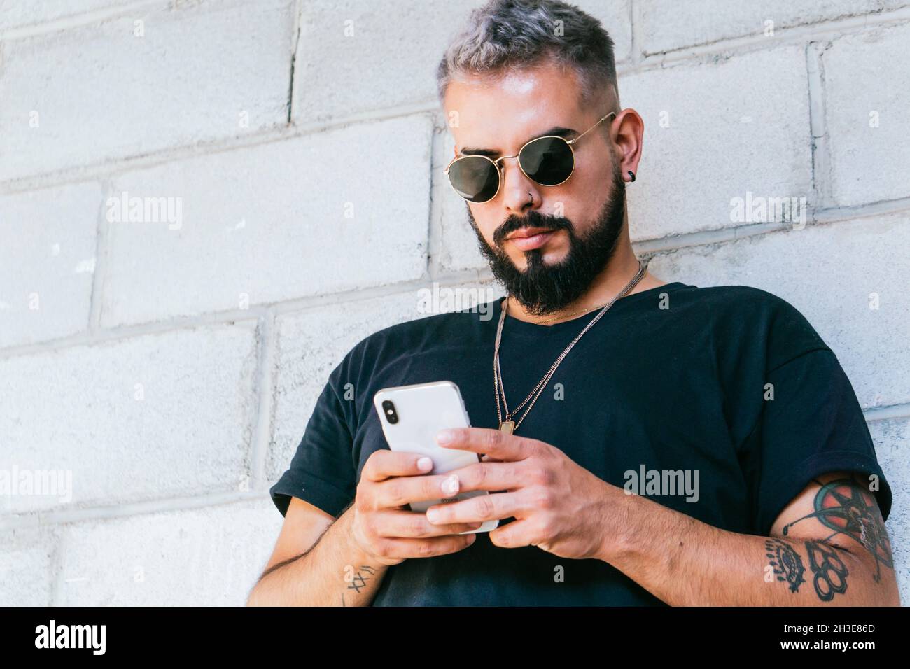 Joyeux barbu avec des tatouages en t-shirt noir et des lunettes de soleil se tenant près du mur du bâtiment et utilisant un smartphone à la lumière du jour Banque D'Images