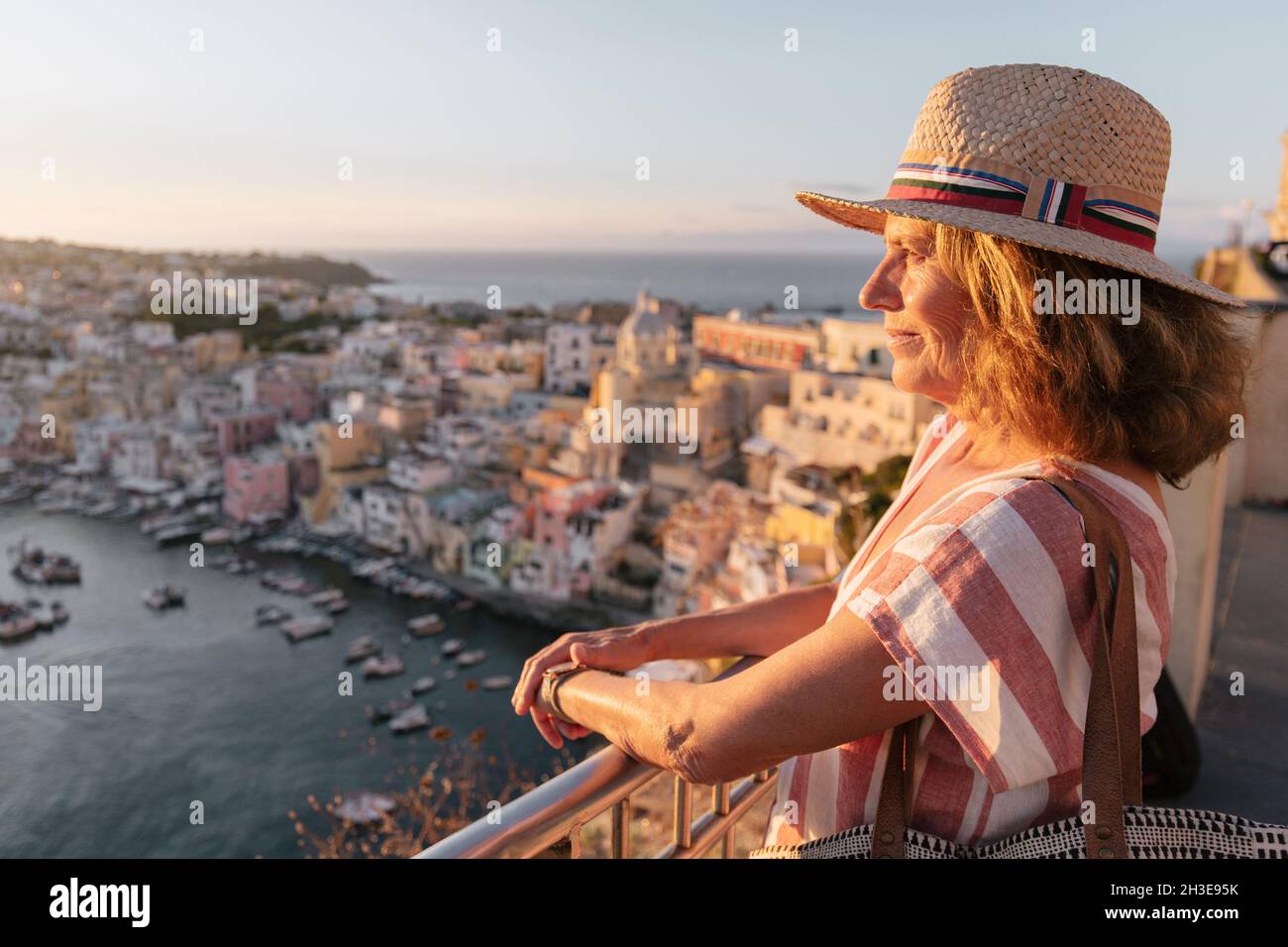 Vue latérale d'une touriste féminine paisible dans une robe élégante et chapeau de paille en admirant la vue pittoresque de l'île de Procida sous le ciel du coucher du soleil Banque D'Images