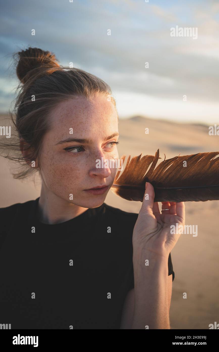 Femme sérieuse avec plume noire portant robe sur dune de sable lavé par la mer au coucher du soleil Banque D'Images