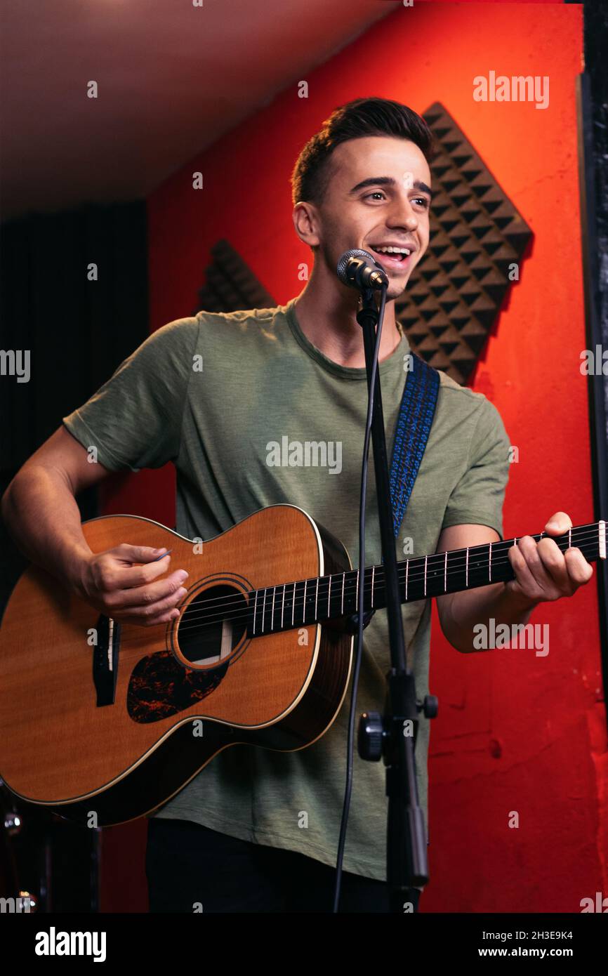 Positif jeune homme guitariste jouant de la guitare acoustique et chantant dans le microphone en club de lumière Banque D'Images