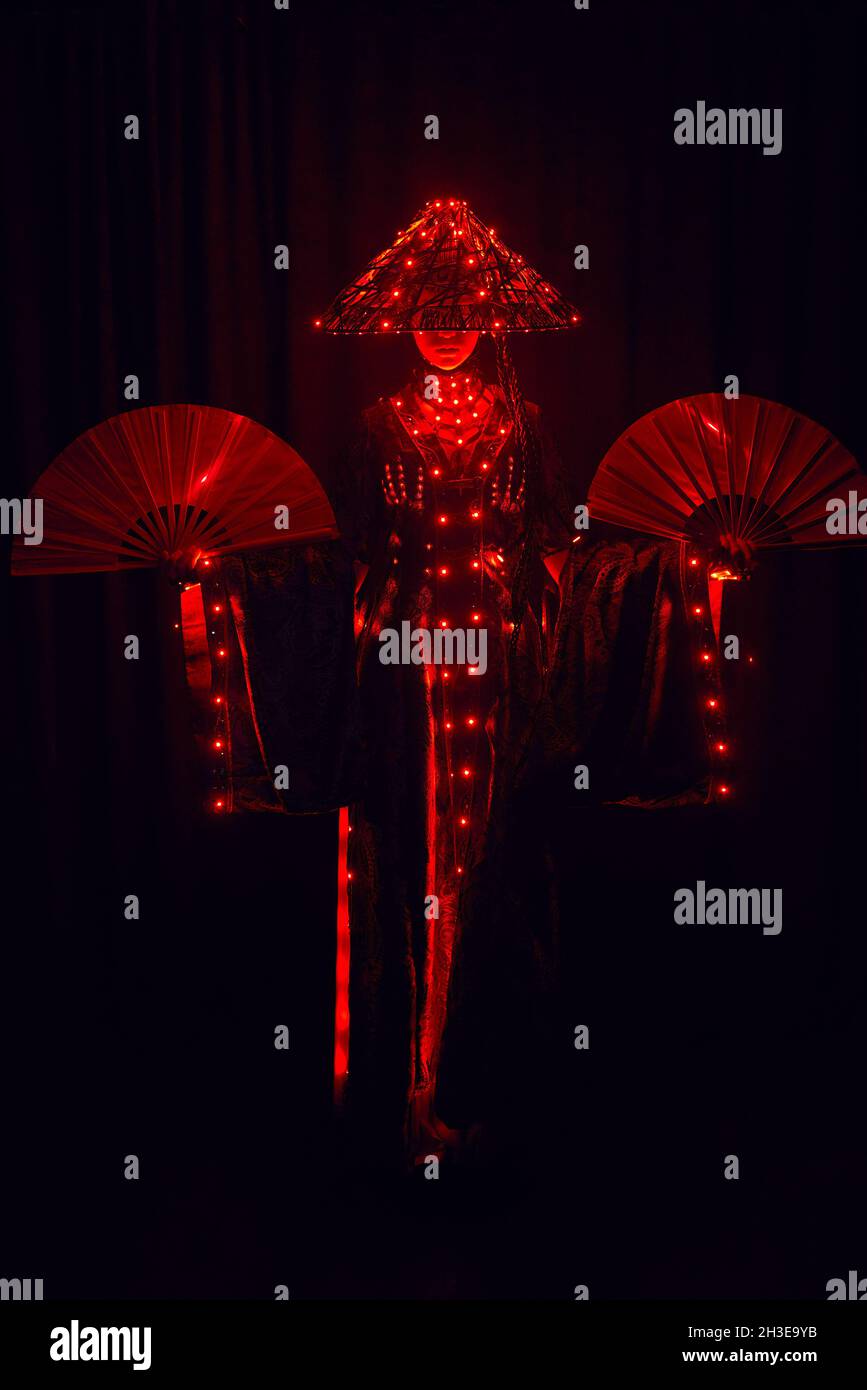 Femme énigmatique méconnaissable dans une tenue traditionnelle créative et coiffeuse vietnamienne avec éclairage rouge debout dans un studio sombre sur fond noir Banque D'Images