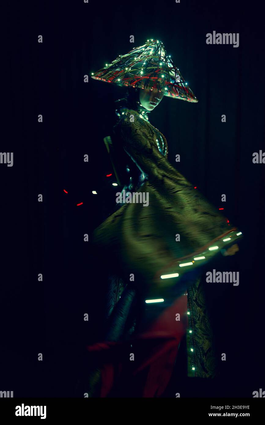 Femme anonyme dans une tenue authentique et chapeau vietnamien avec éclairage lumineux debout dans un studio moderne sombre sur fond noir pendant les performances Banque D'Images