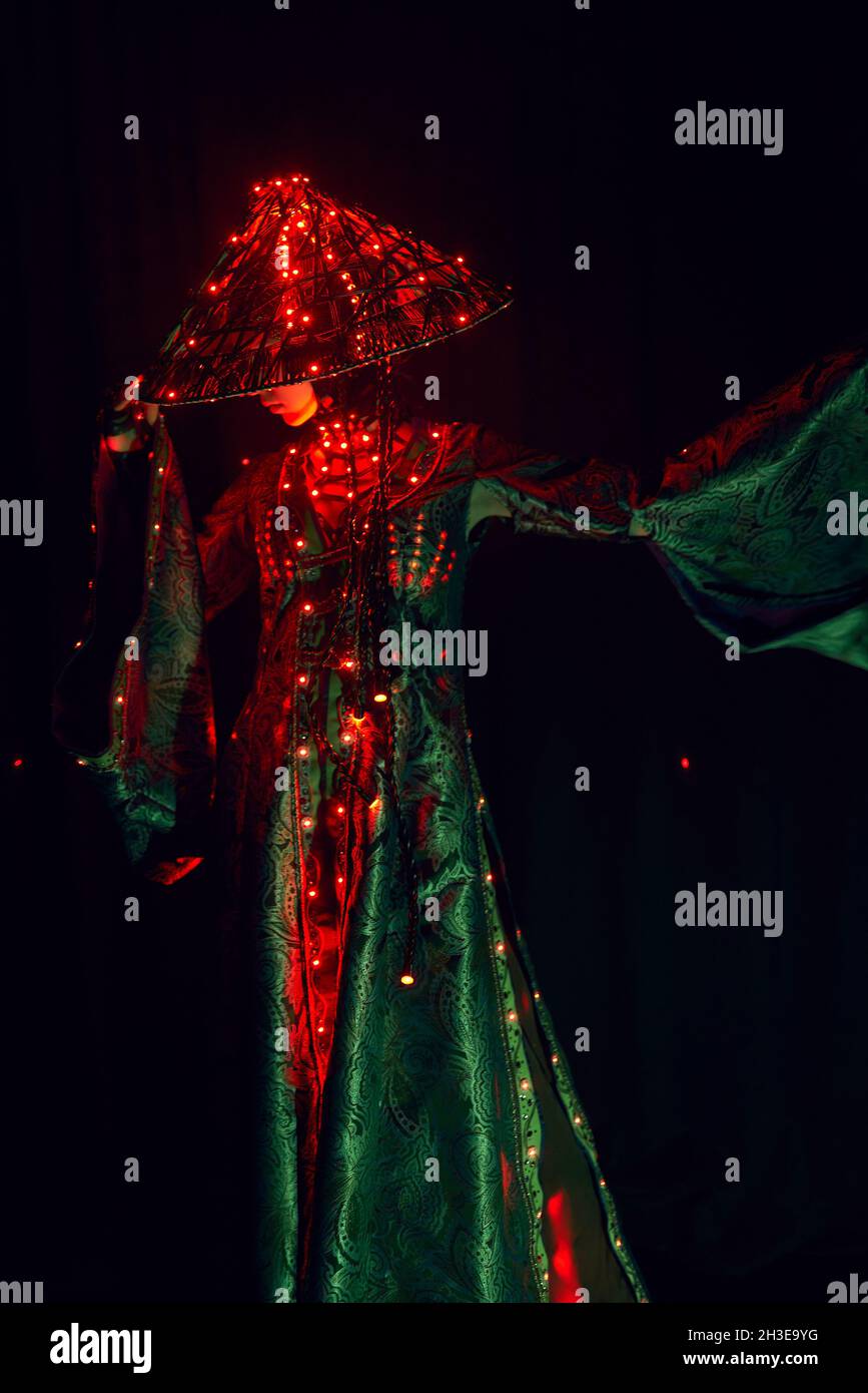 Femme énigmatique méconnaissable dans une tenue traditionnelle créative et coiffeuse vietnamienne avec éclairage rouge debout dans un studio sombre sur fond noir Banque D'Images