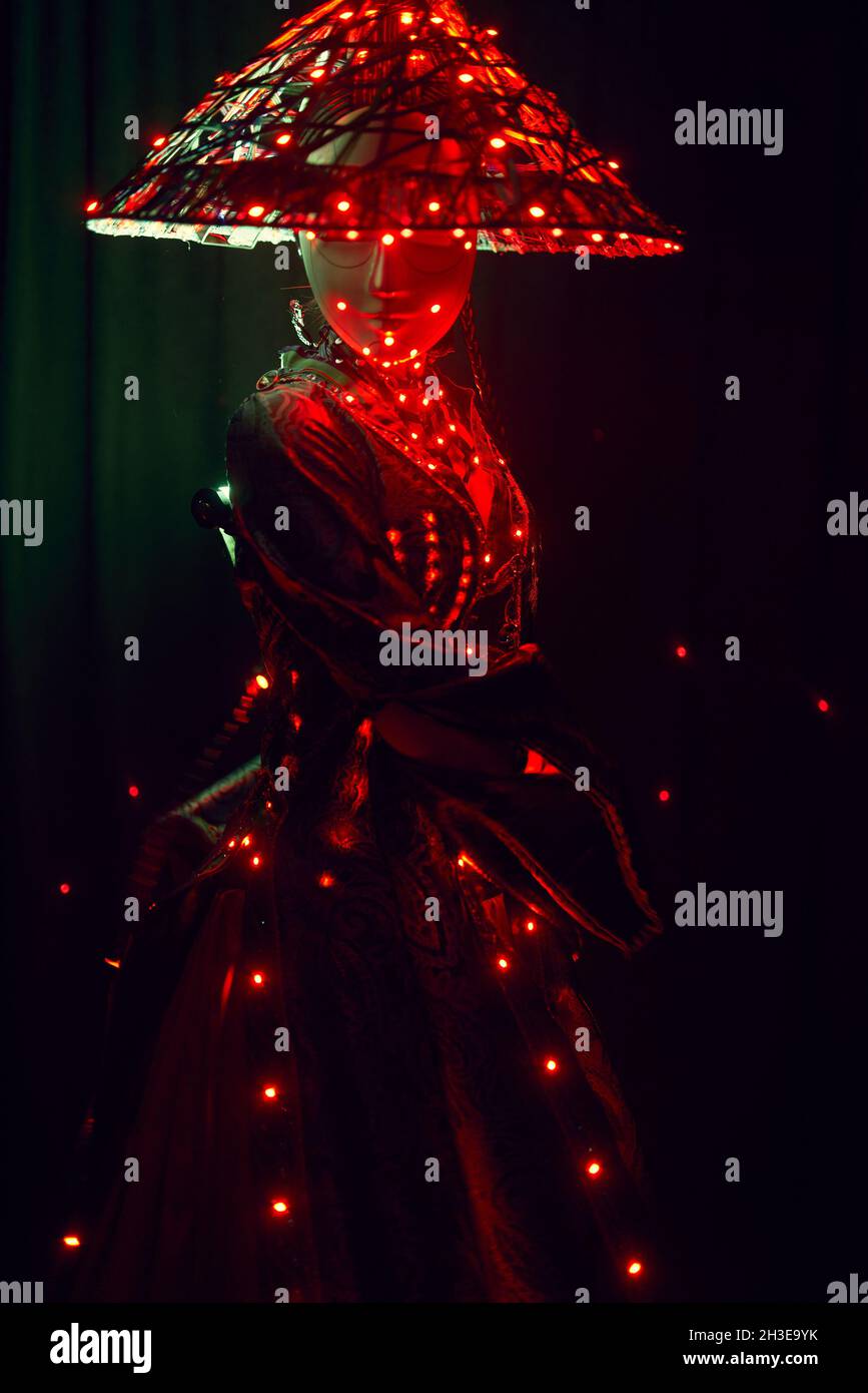 Femme énigmatique méconnaissable dans une tenue traditionnelle créative et coiffeuse vietnamienne avec éclairage rouge debout dans un studio sombre sur fond noir Banque D'Images