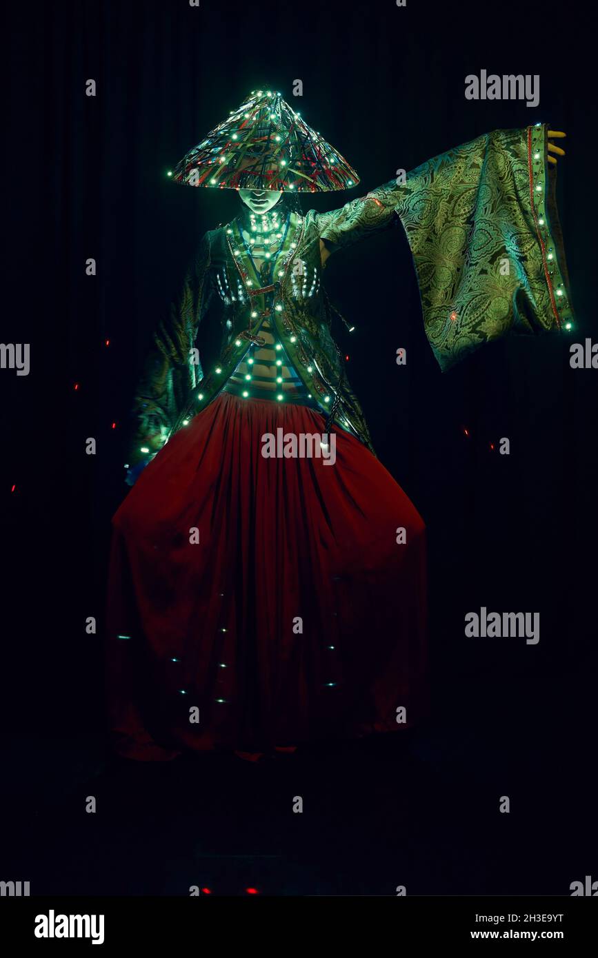 Femme anonyme dans une tenue authentique et chapeau vietnamien avec éclairage lumineux debout dans un studio moderne sombre sur fond noir pendant les performances Banque D'Images