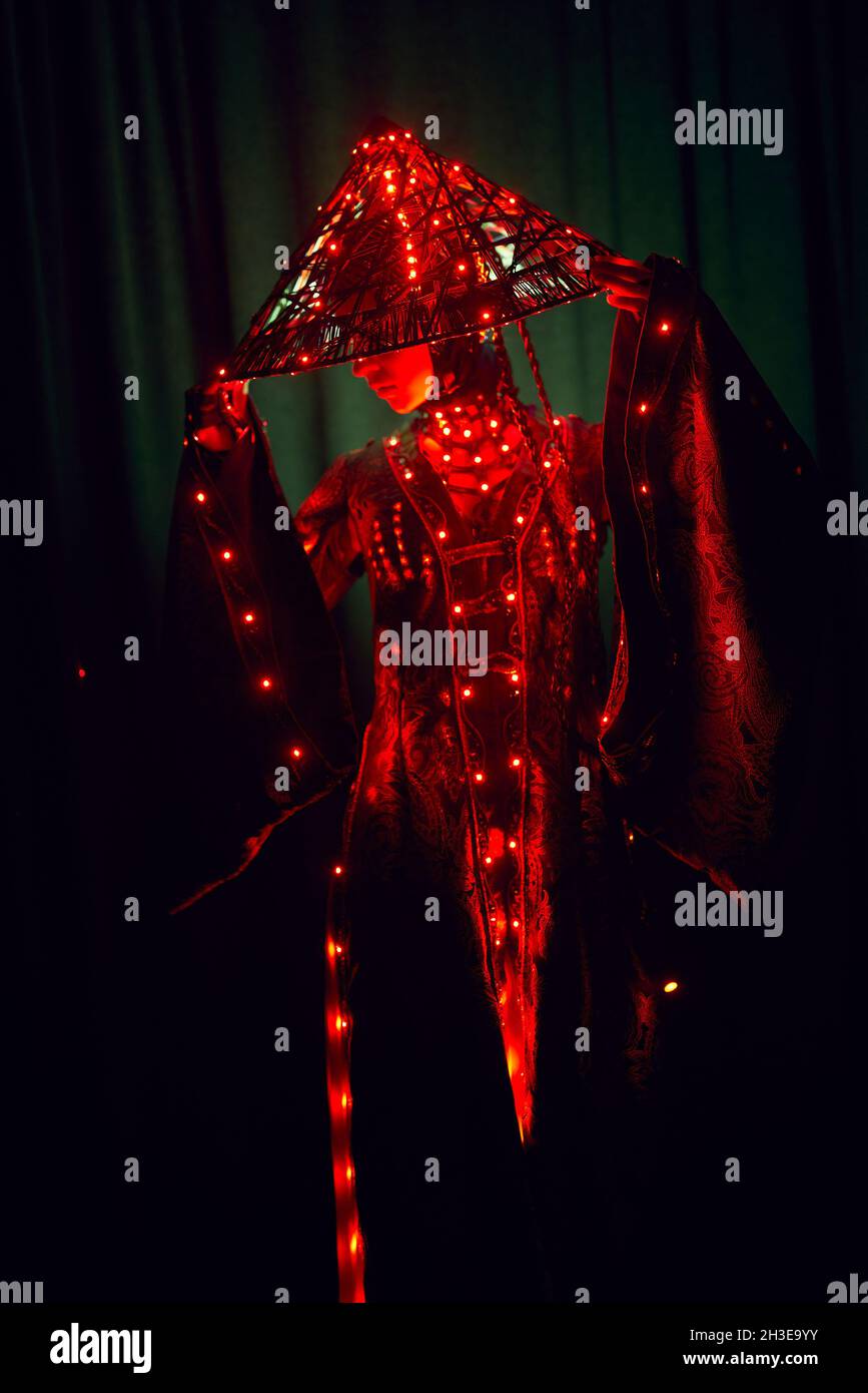 Femme énigmatique méconnaissable dans une tenue traditionnelle créative et coiffeuse vietnamienne avec éclairage rouge debout dans un studio sombre sur fond noir Banque D'Images