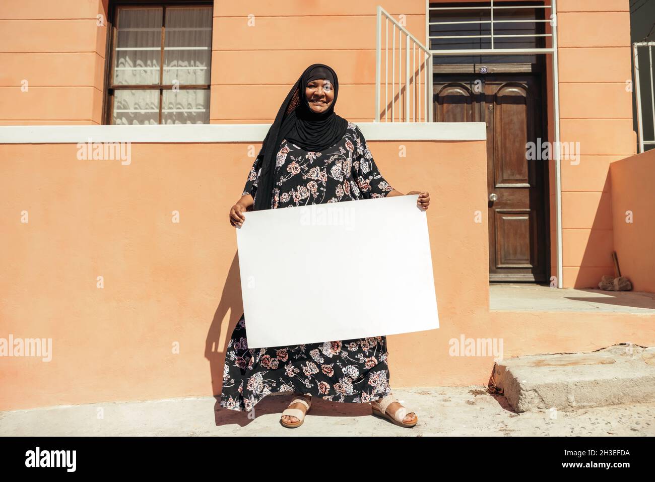 Bonne femme musulmane tenant une affiche blanche et souriant à la caméra tout en se tenant devant sa maison pendant la journée.Confiance en soi, l'homme d'âge moyen Banque D'Images Bonne femme musulmane tenant une affiche blanche et souriant à la caméra tout en se tenant devant sa maison pendant la journée.Confiance en soi, l'homme d'âge moyen Banque D'Images