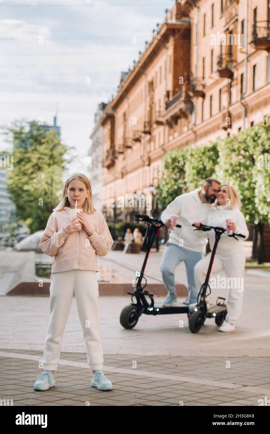 Une fille avec une boisson dans ses mains est debout dans la ville et derrière elle sur des scooters électriques sont ses parents. Banque D'Images