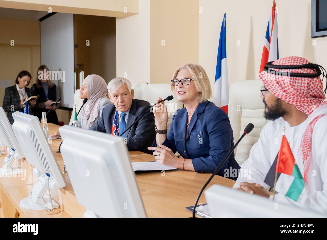 Femme politique confiante et mature en lunettes assise à la table de conférence et parlant au micro lors du congrès national Banque D'Images