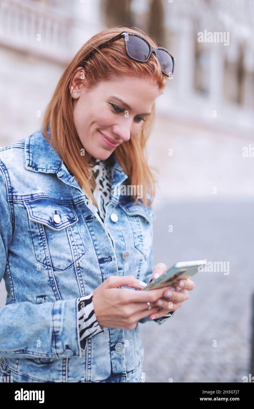 Jeune femme caucasienne à tête rouge messagerie sur smartphone à l'extérieur, verticale Banque D'Images