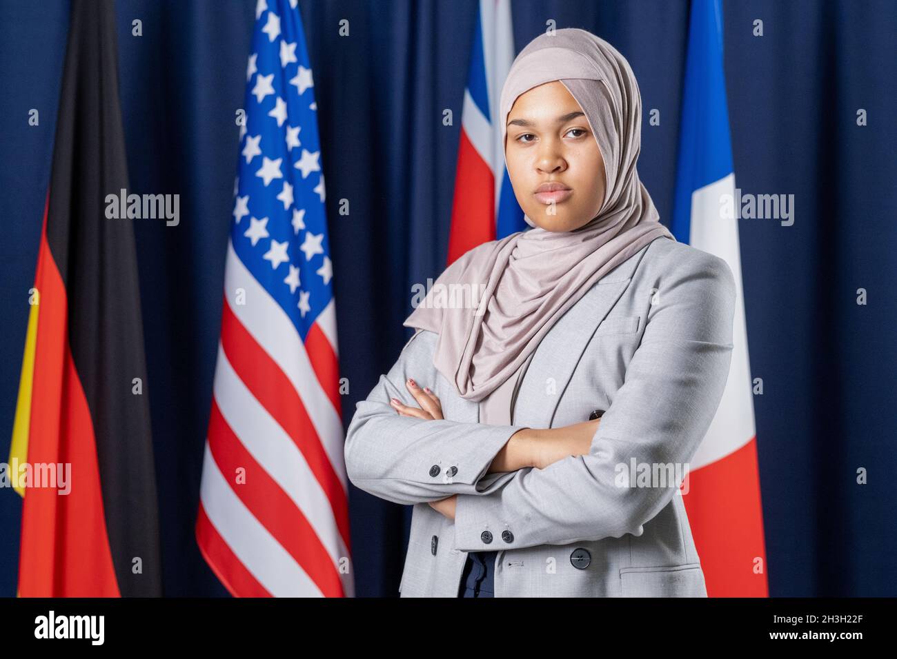 Portrait d’une femme musulmane sérieuse et confiante en veste officielle et hijab debout avec des armes croisées contre des drapeaux nationaux Banque D'Images