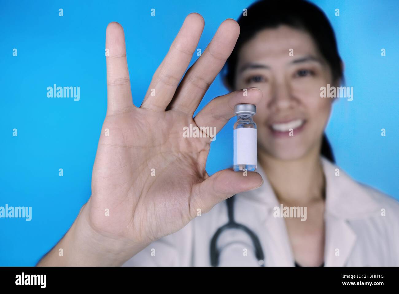 Un médecin attrayant, portant un manteau blanc avec un stéthoscope autour de son épaule, tenant un flacon en verre de flacon de vaccin avec une étiquette blanche vierge, smili Banque D'Images