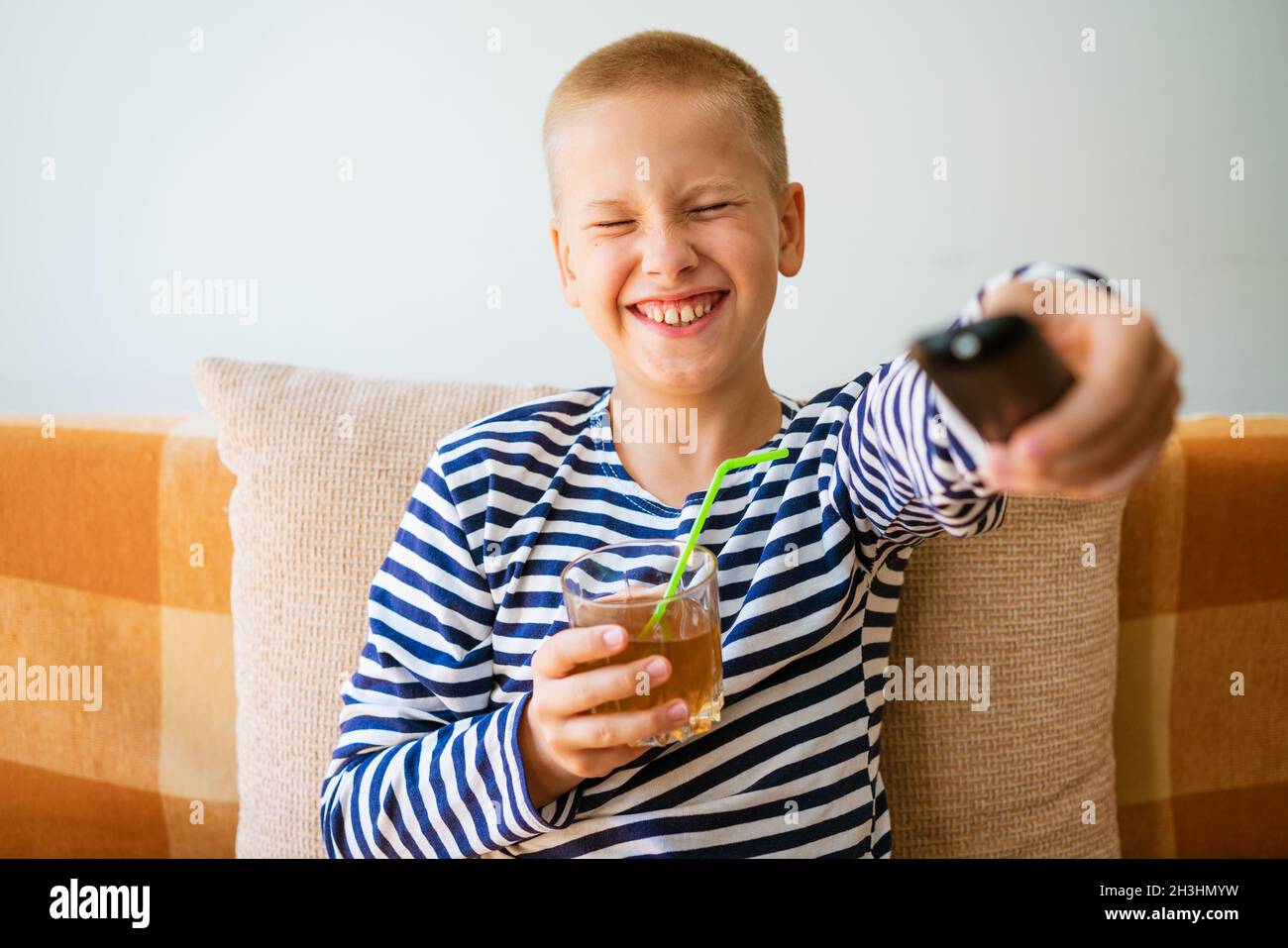 Un garçon d'âge scolaire caucasien est assis sur un canapé dans des vêtements décontractés avec une télécommande dans sa main et change de chaîne sur la télévision. Et boire du jus dans un verre à travers une paille. Concept de loisirs maison de vacances Banque D'Images
