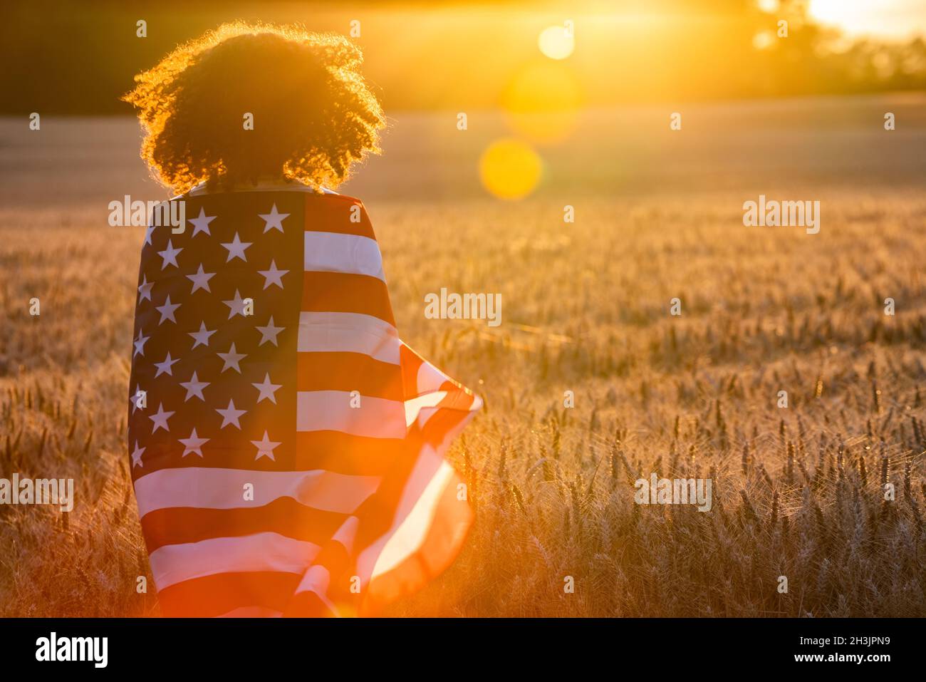 Mixed Race African American girl adolescent femme jeune femme dans un champ de blé ou d'orge enveloppés dans USA stars and stripes flag dans golden sunse Banque D'Images