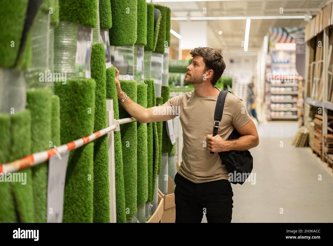 Un gars décontracté pensif en magasin avec des articles de maison choisissant l'herbe artificielle dans les rouleaux exposés Banque D'Images
