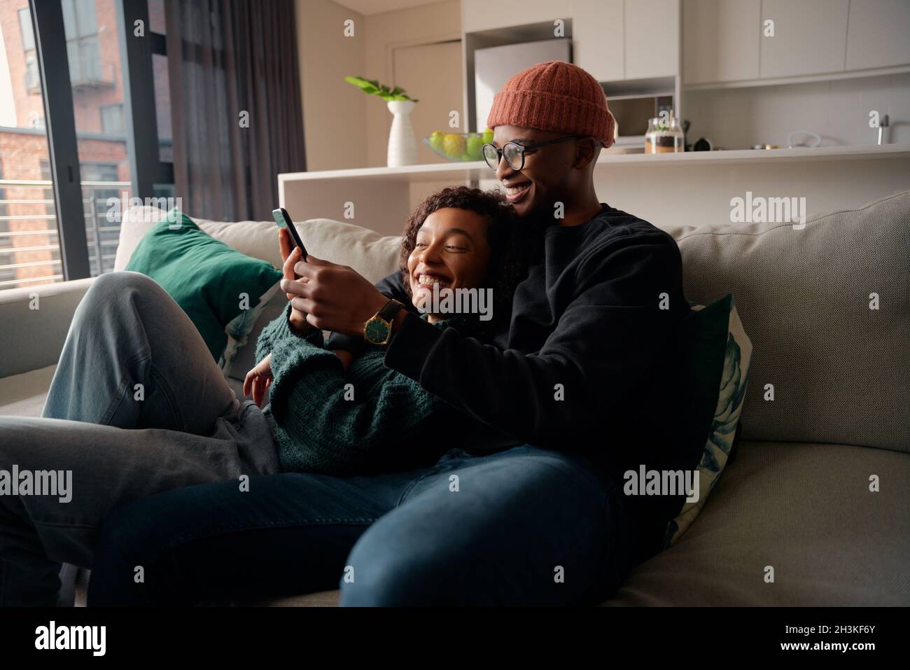 Un couple multiethnique riant au téléphone tout en étant sur le canapé dans un appartement moderne Banque D'Images