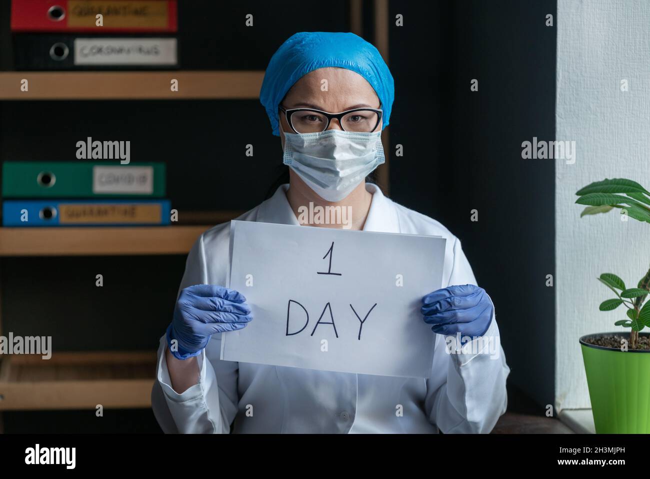 Médecin avec des signes UN JOUR debout près de la fenêtre dans le bureau médical.Le port de l'uniforme et du masque de protection médic féminin montre le quarant Banque D'Images