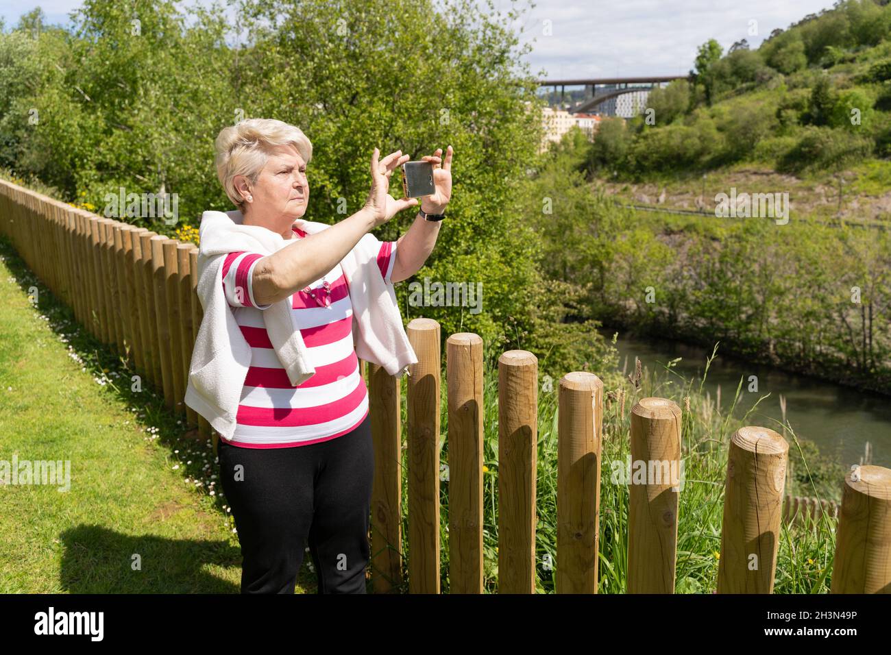 Femme âgée faisant de la vidéo panoramique avec téléphone au parc le jour ensoleillé.Femme aînée debout près d'une clôture, prenant des photos avec un smartphone sur un paysage naturel Banque D'Images