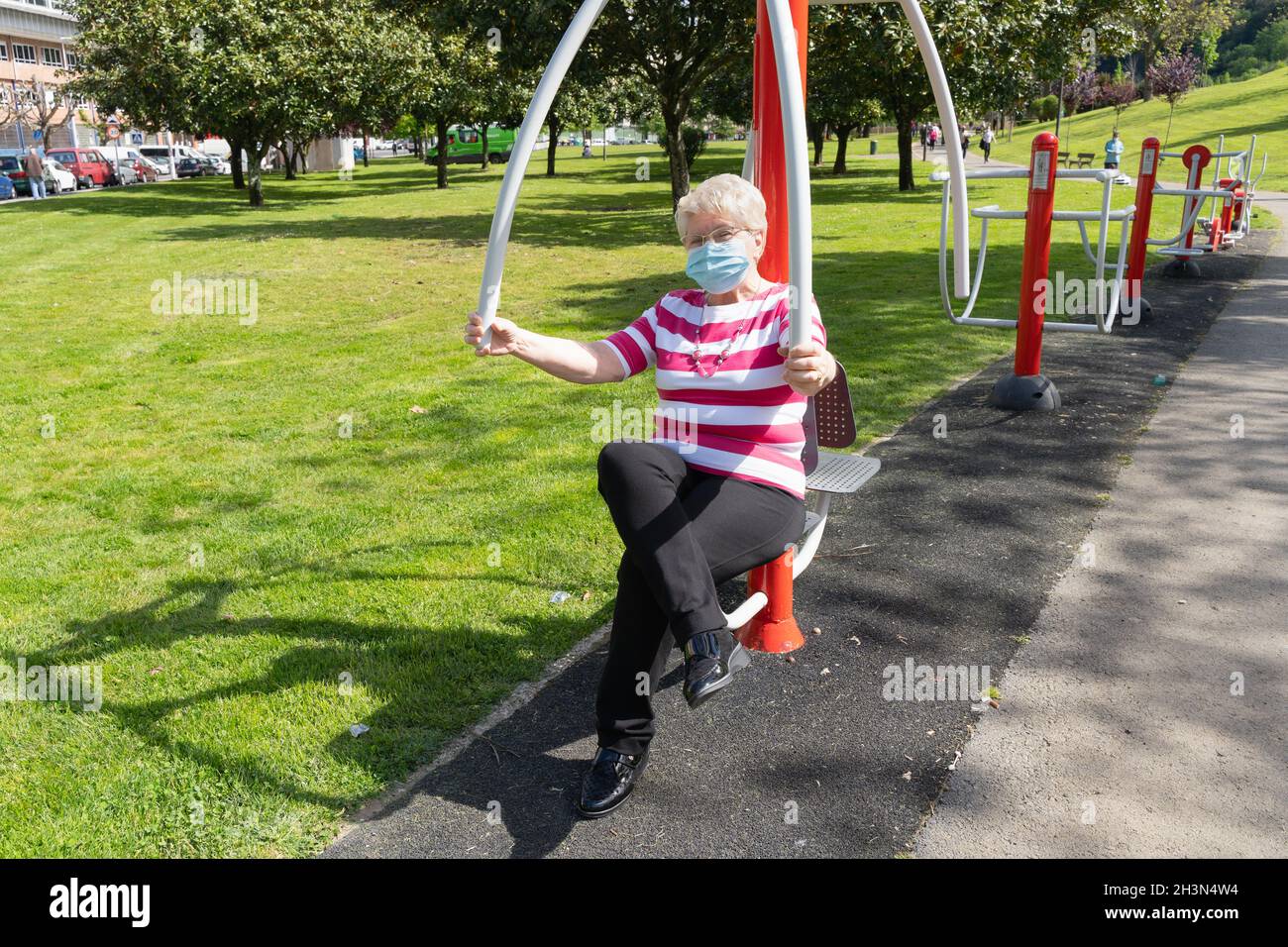 Femme blonde aînée avec masque de visage faisant de l'exercice sur la machine du parc le jour ensoleillé.Femme senior s'entraîne dans le haut du corps pour garder la forme et la santé à l'extérieur Banque D'Images
