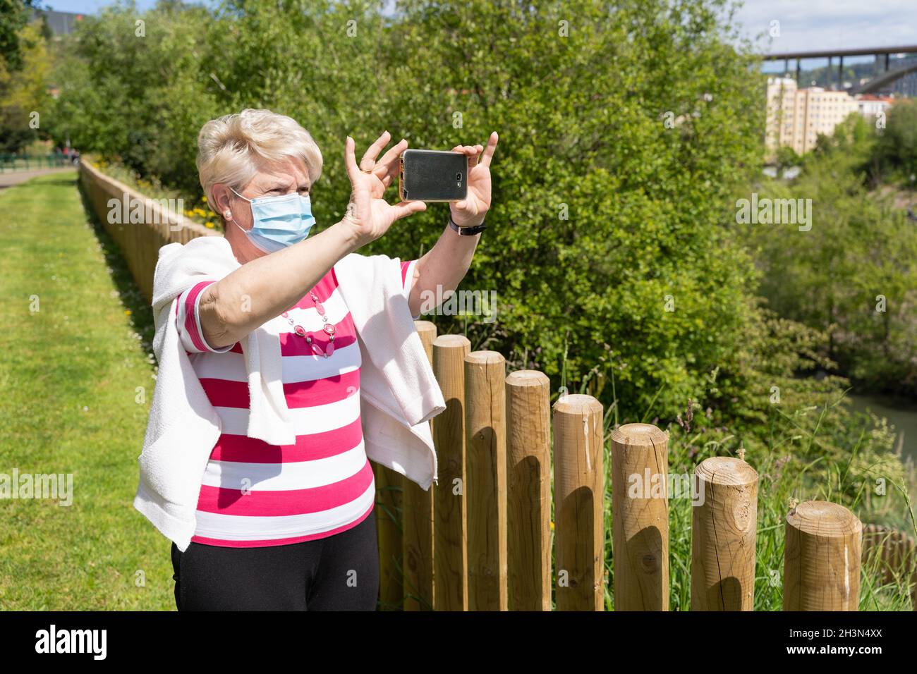 Femme âgée avec masque de visage faisant de la vidéo panoramique avec téléphone portable au parc le jour ensoleillé.Femme aînée debout près d'une clôture, prenant des photos avec un smartphone Banque D'Images