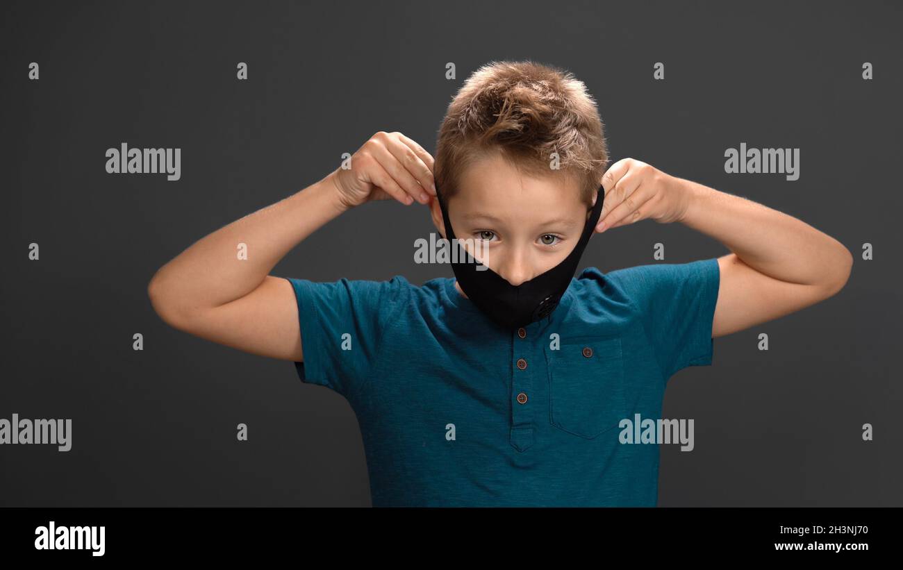 Petit garçon prend son masque noir avec sa main sur fond gris.L'enfant caucasien respire avec soulagement et sourires regardant Banque D'Images